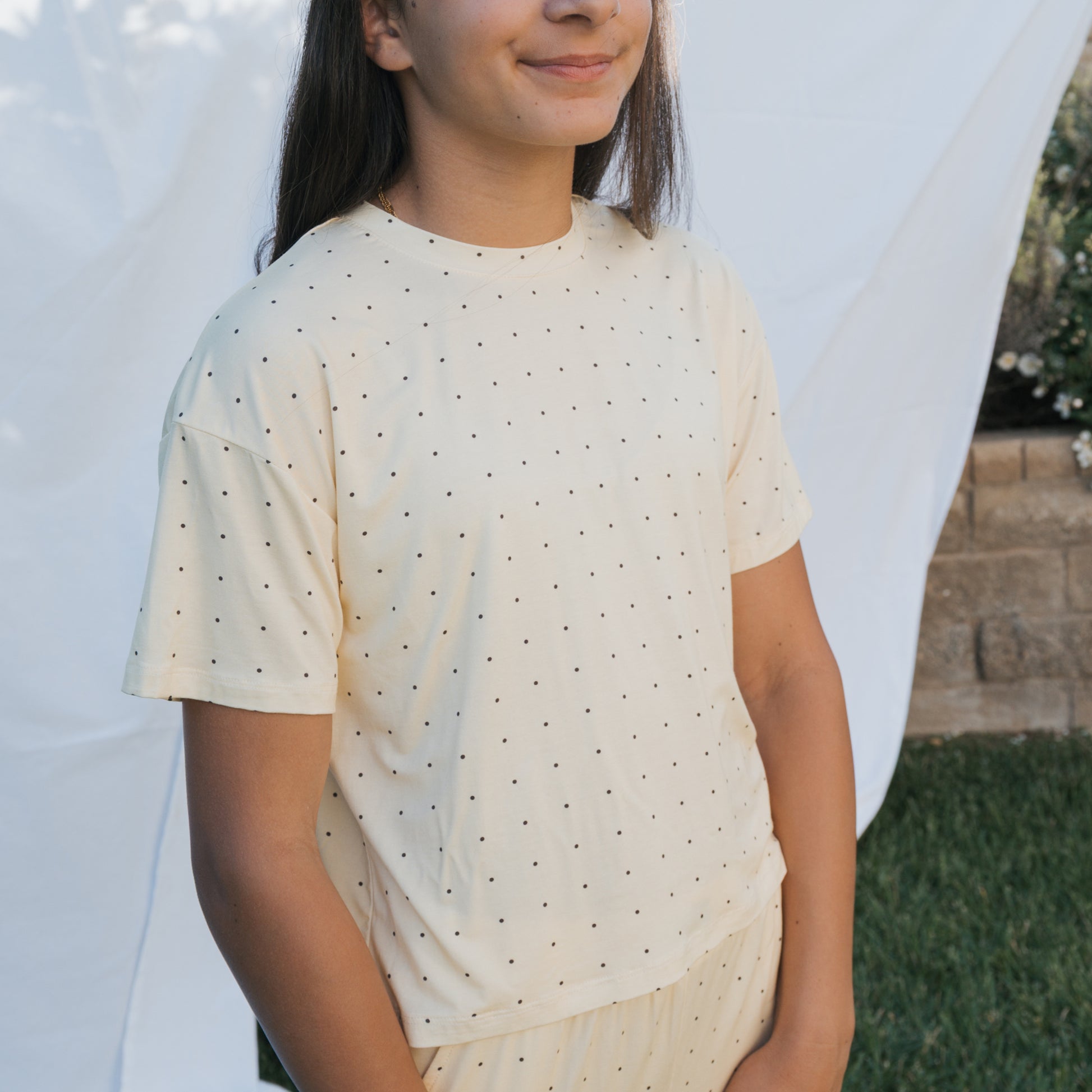 A person stands outdoors in front of a white sheet backdrop, wearing the forever french baby Child Boxy Tee Lounge Set | Veda. They have long dark hair, are smiling slightly with hands in front. Grass and a stone wall are visible.
