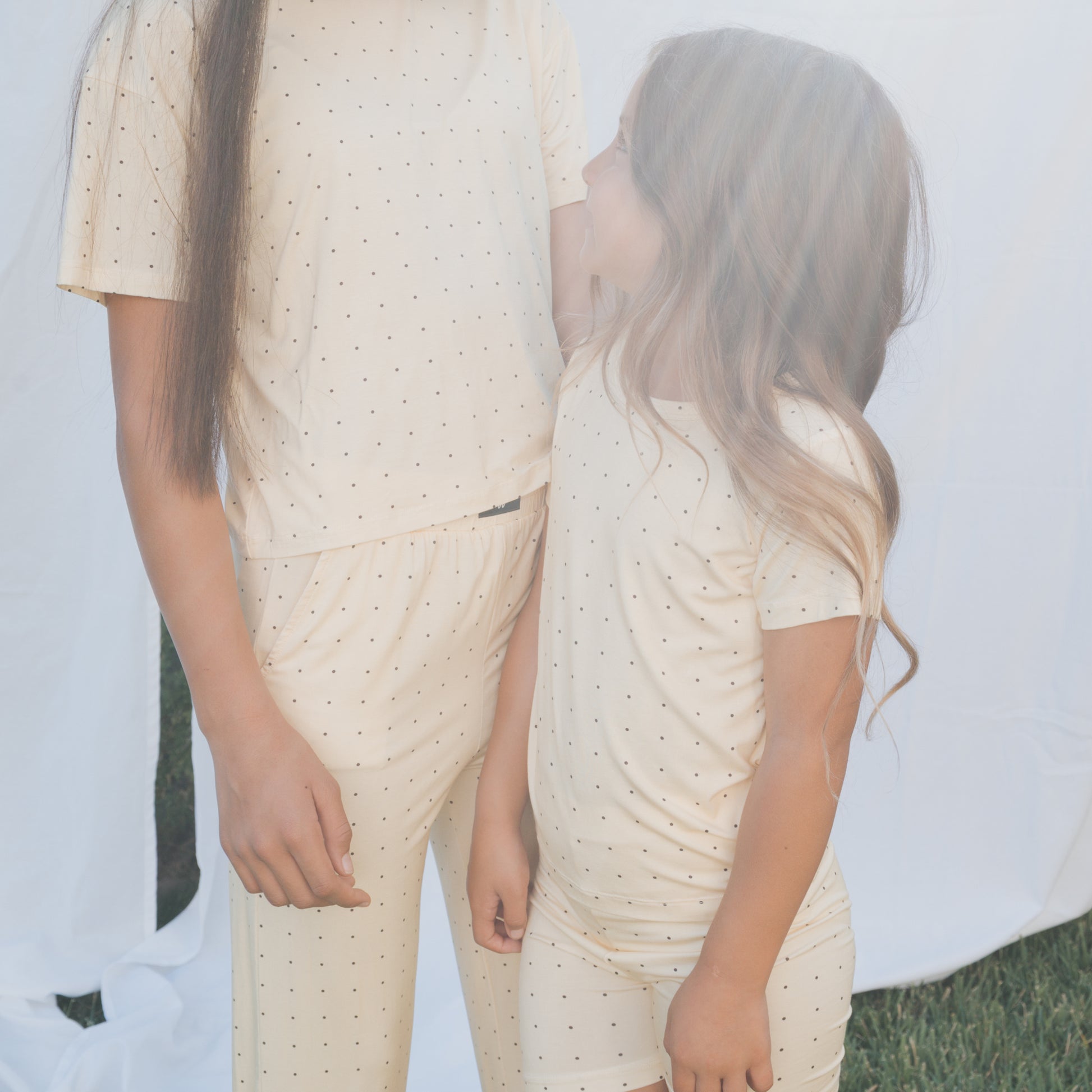 A child in the forever french baby Child Boxy Tee Lounge Set | Veda stands outdoors with an adult, both in soft bamboo clothing with subtle dots. White fabric drapes in the background, creating a gentle, hypo-allergenic mood.
