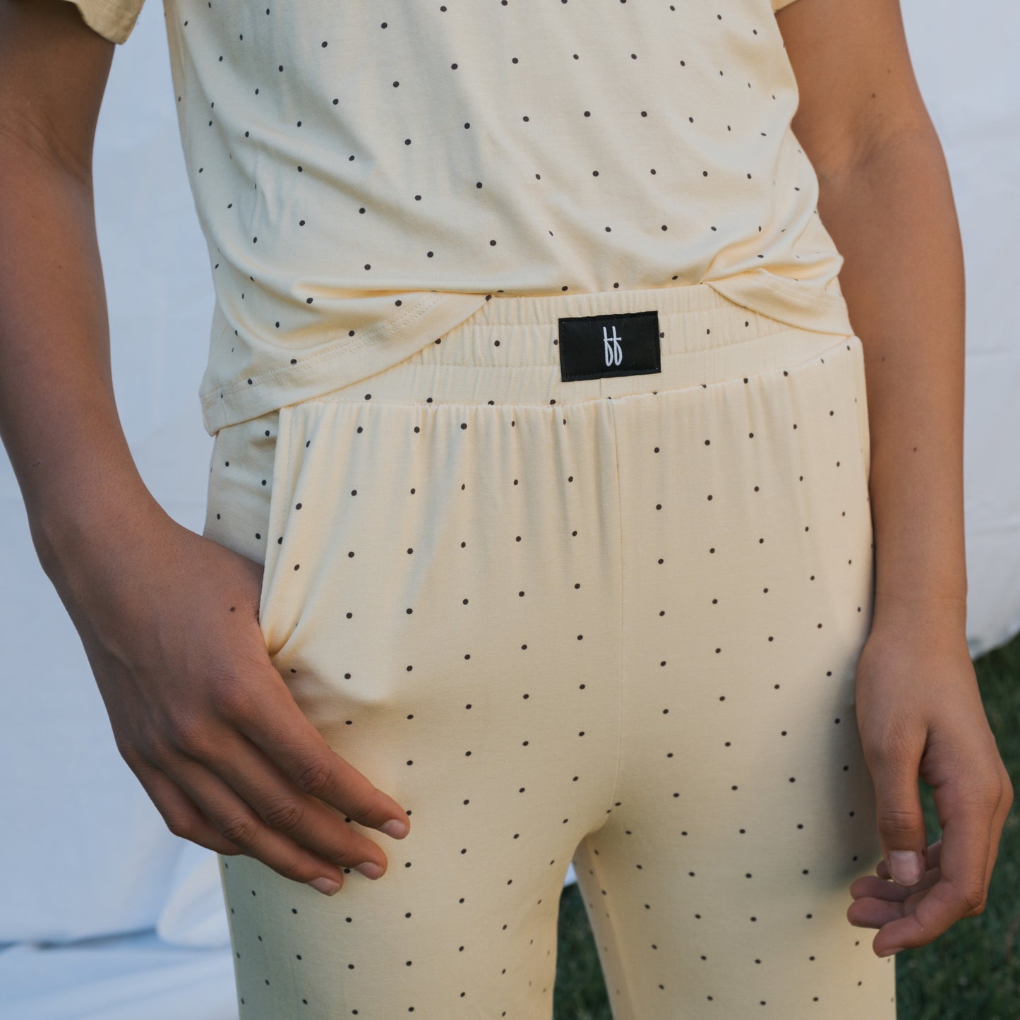 A child wears the forever french baby Child Boxy Tee Lounge Set | Veda in light yellow with black polka dots outdoors, hands near the waistband featuring a black tag with white lettering.
