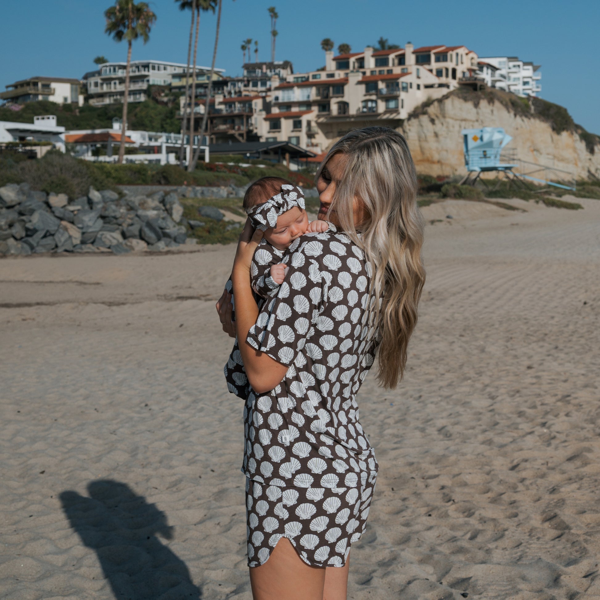 A woman holding a baby on a sandy beach, both in matching forever french baby Women's Bamboo Short Set | Seashells, with beach houses and palm trees in the background beneath the clear blue sky.