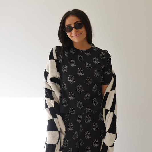 A woman with long dark hair in sunglasses wears the forever french baby Women's Boxy Tee Lounge Set | Flaming Hearts, featuring a black tee and white abstract pants, with a checkered sweater over her shoulders, smiling against a plain light background.