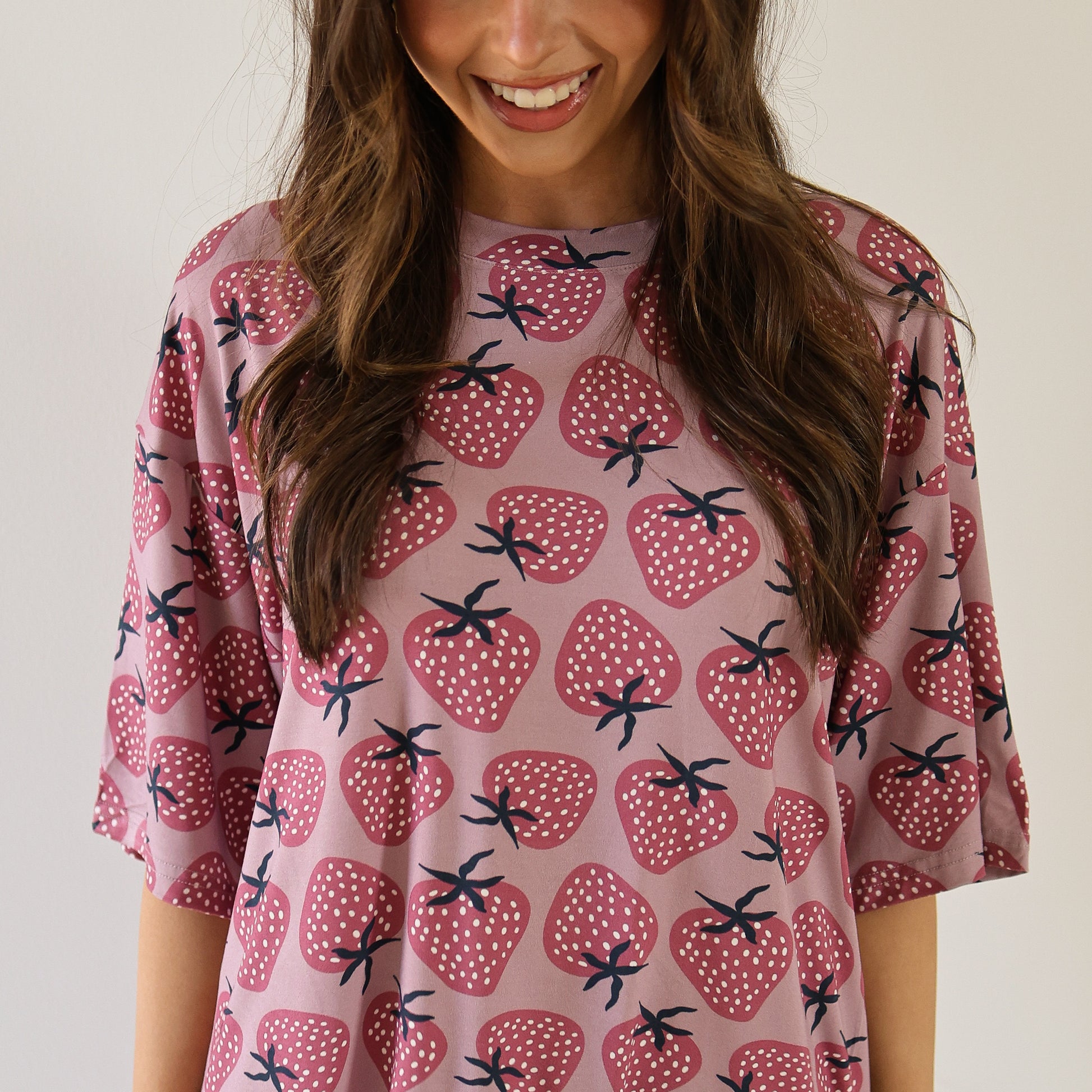 A woman with long brown hair smiles while wearing the forever french baby Women's Bamboo Short Set | Matilda, featuring a light purple top with large red strawberries and green stems. The background is plain white.