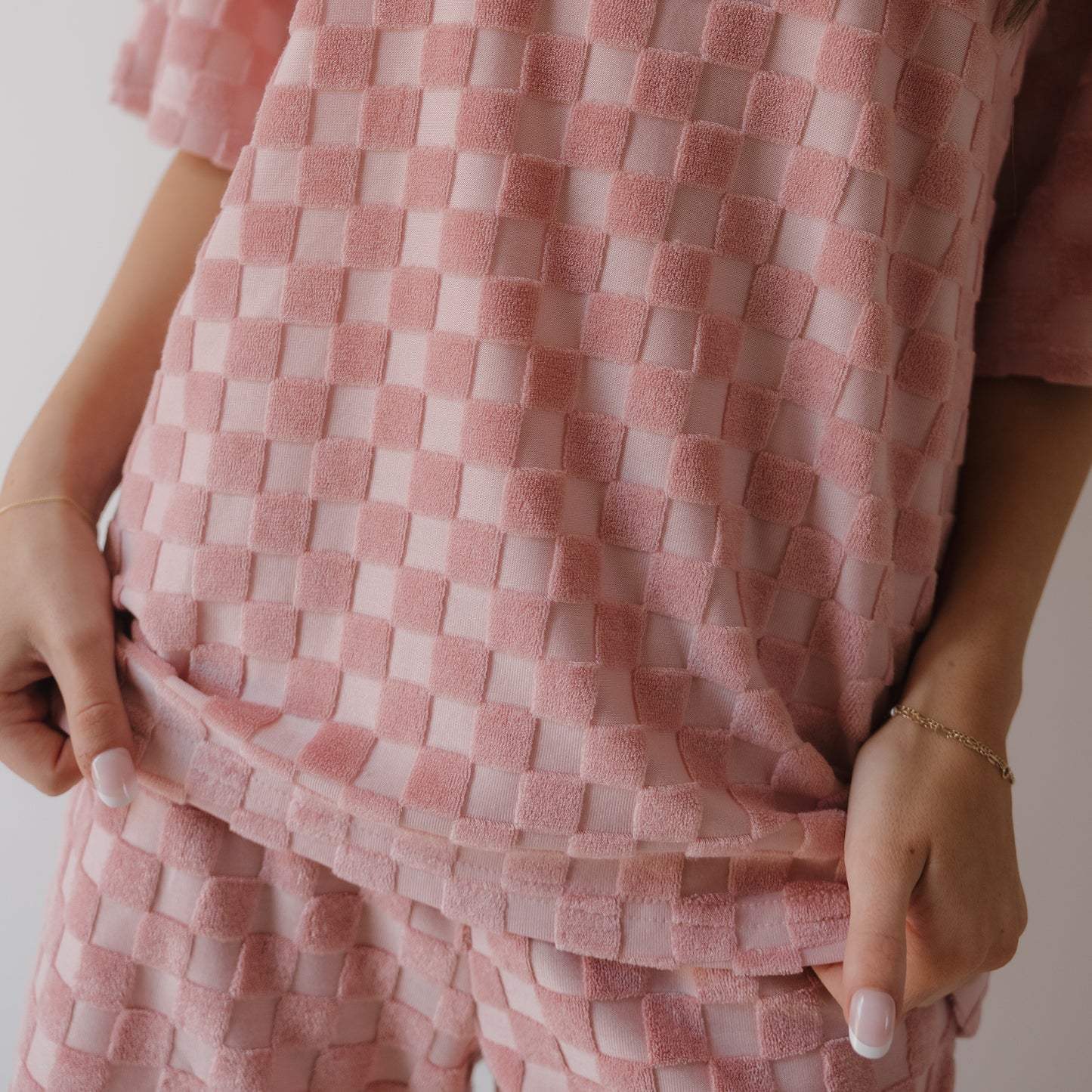 A woman models the forever french baby Women's Terry Short Set in Sea Pink, gently holding the hem of her matching shirt to showcase the coordinating shorts. Only her lower torso and hands are visible.