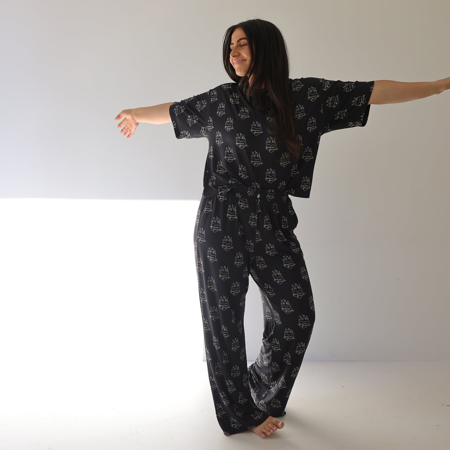 A woman with long dark hair stands barefoot, smiling gently in a bright, minimal room. She wears the forever french baby Women's Boxy Tee Lounge Set in the Flaming Hearts pattern with arms outstretched.