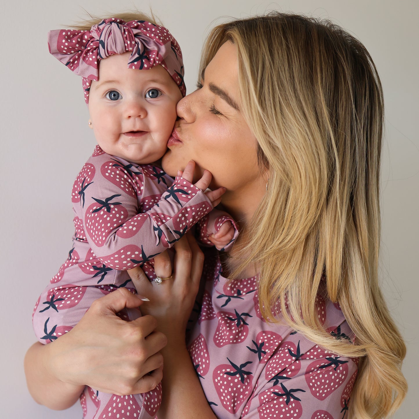 A woman with long blonde hair kisses a baby in matching "Bamboo Zip Pajamas | Matilda" by forever french baby. The strawberry-patterned pink outfits include a bow headband for the baby. They stand against a plain background.