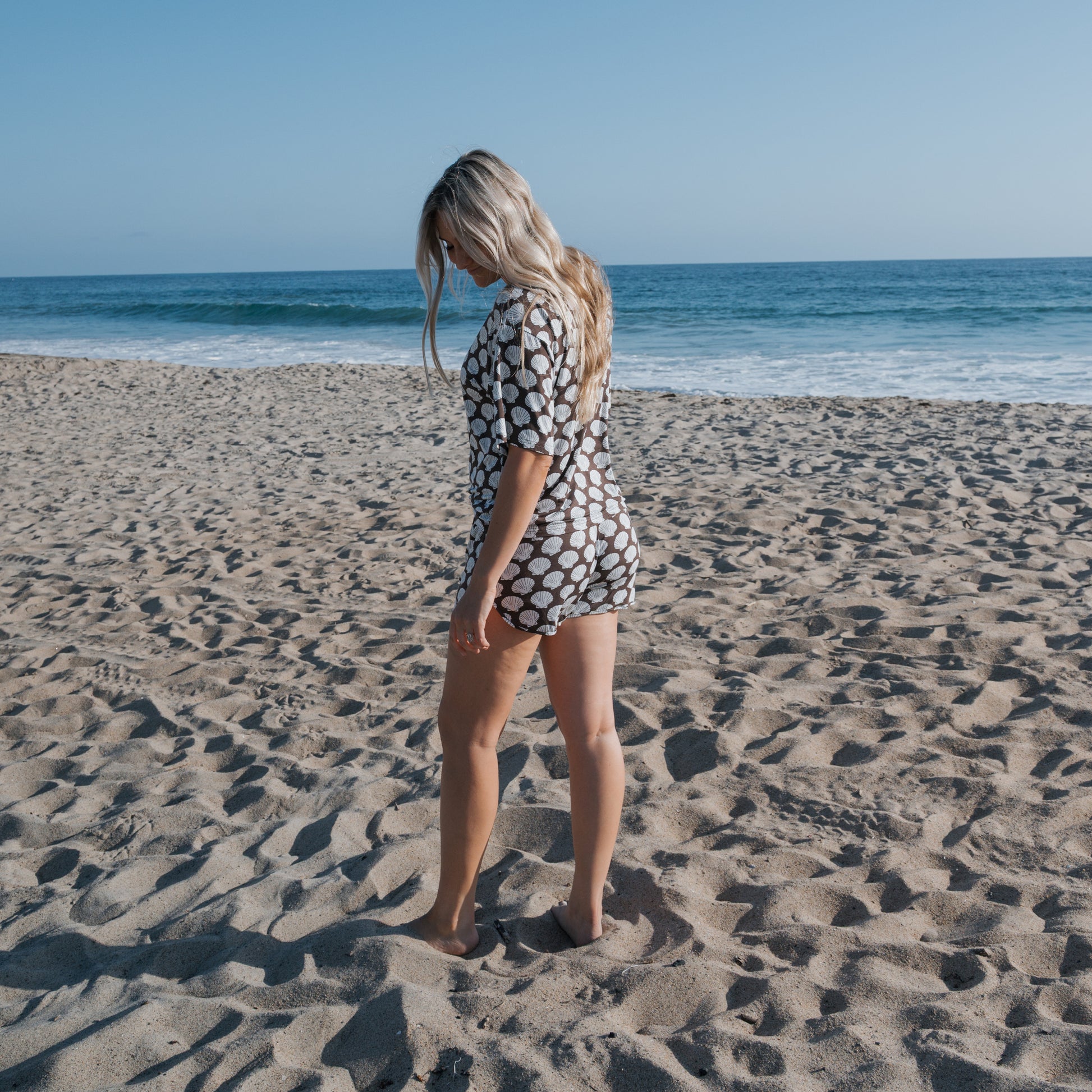 A woman with long blonde hair wears the Women's Bamboo Short Set | Seashells by forever french baby, standing barefoot on a sandy beach with the ocean and blue sky in the background.