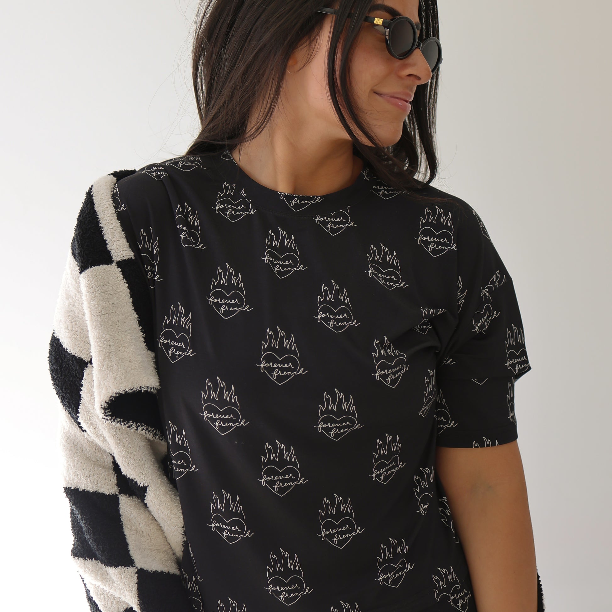 Woman with long dark hair wearing black sunglasses and the Women's Boxy Tee Lounge Set | Flaming Hearts by forever french baby, featuring a white flame and lips design, with a checkered jacket over her shoulder against a light background.
