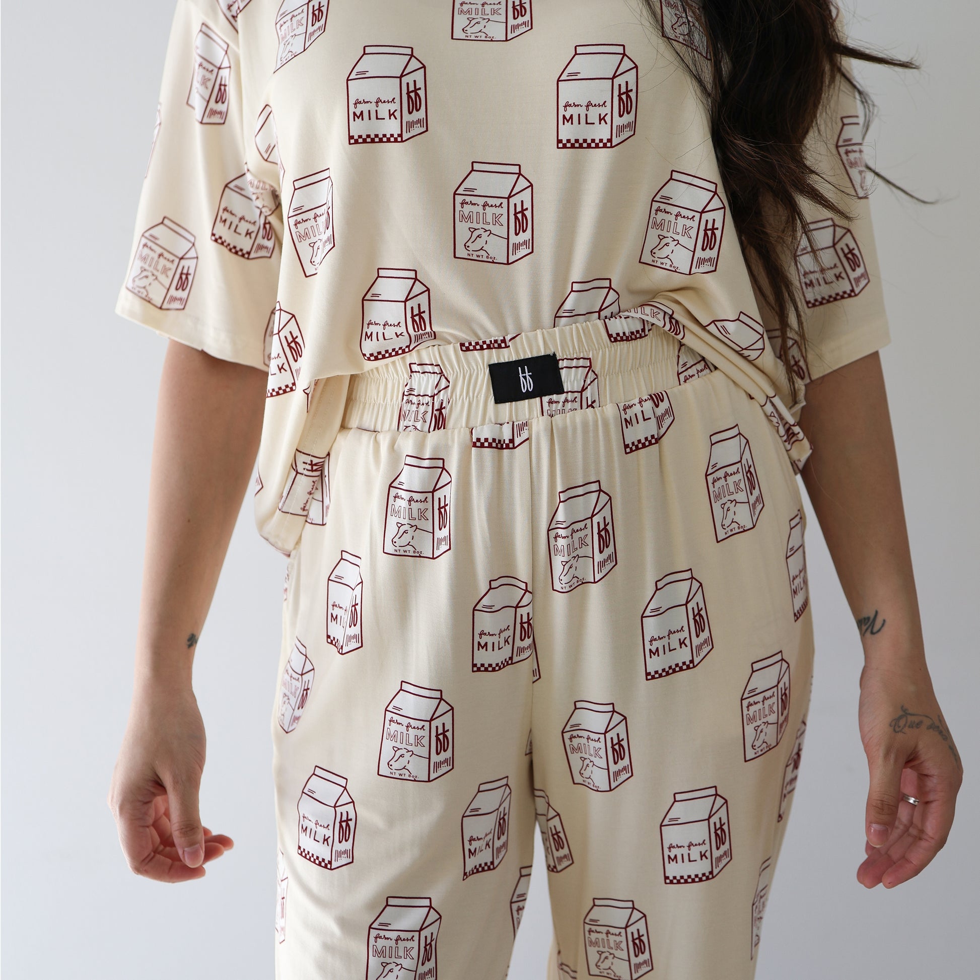 A person wears the forever french baby Women's Boxy Tee Lounge Set | Milk in cream with red milk carton illustrations. Only their torso and upper legs show, one arm hangs down displaying faint forearm and hand tattoos.