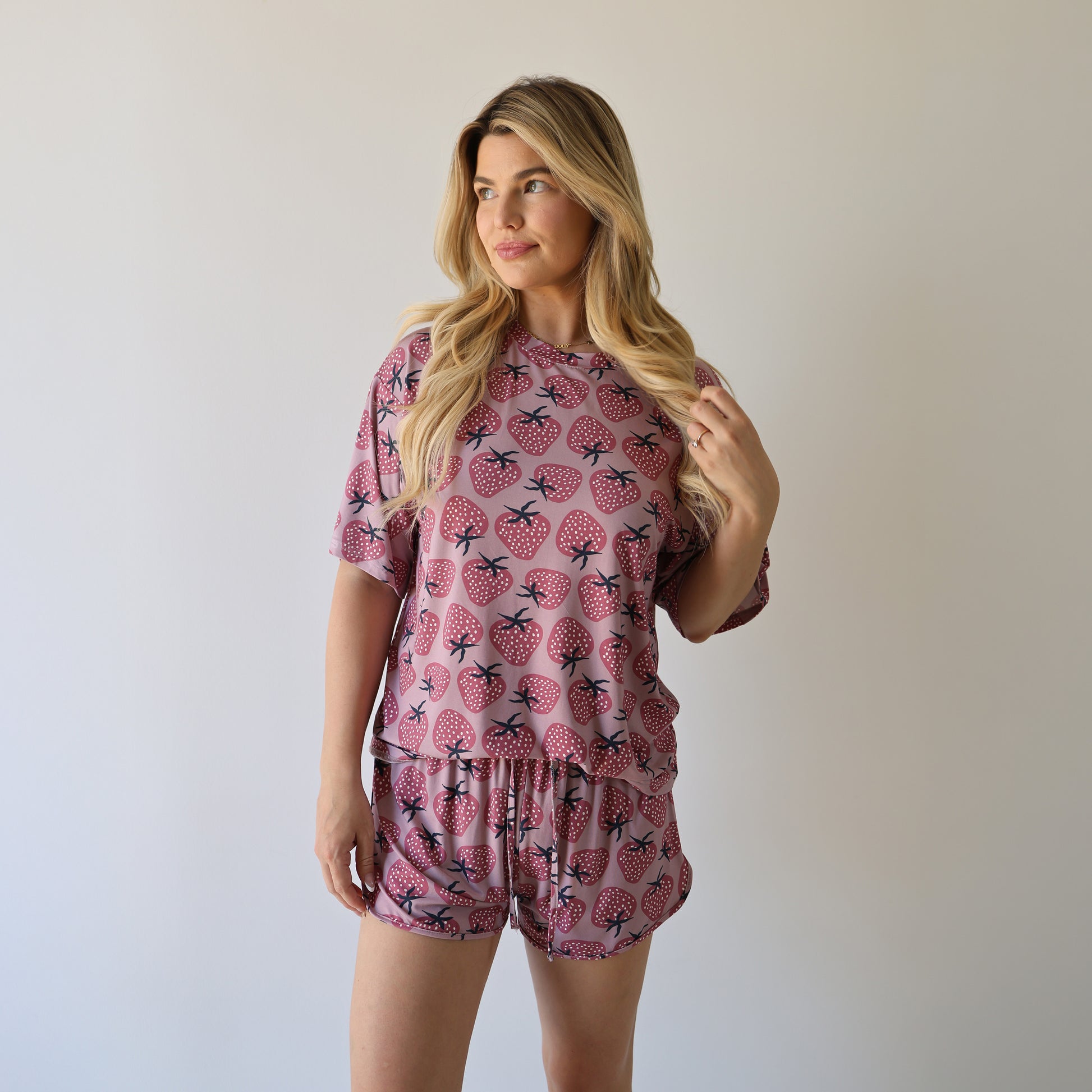 A woman with long blonde hair wears the forever french baby Women's Bamboo Short Set | Matilda, featuring a pink strawberry print. She stands against a light background, gazing sideways with one hand in her hair.