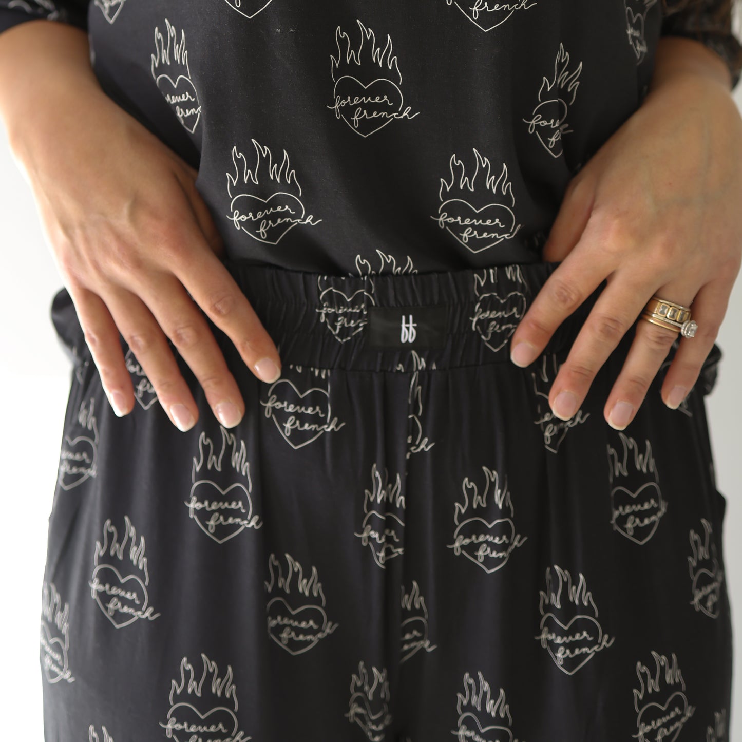 A person wears the forever french baby Women's Boxy Tee Lounge Set | Flaming Hearts, featuring matching black loungewear with flaming heart graphics, styled with a comfortable, hypo-allergenic gold ring for everyday wear.