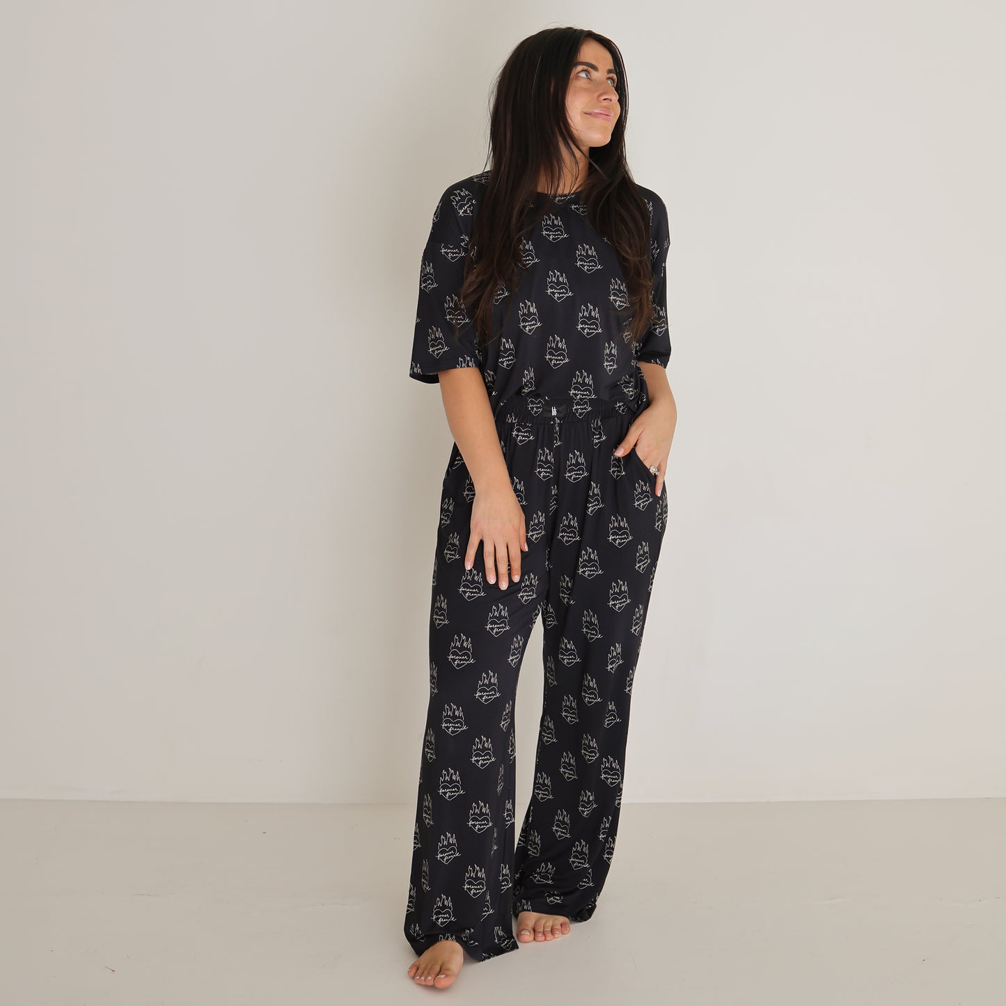 A woman with long, dark hair smiles as she stands barefoot against a white background, wearing the forever french baby Women's Boxy Tee Lounge Set | Flaming Hearts—a dark, loose-fitting bamboo loungewear set with a white abstract pattern.