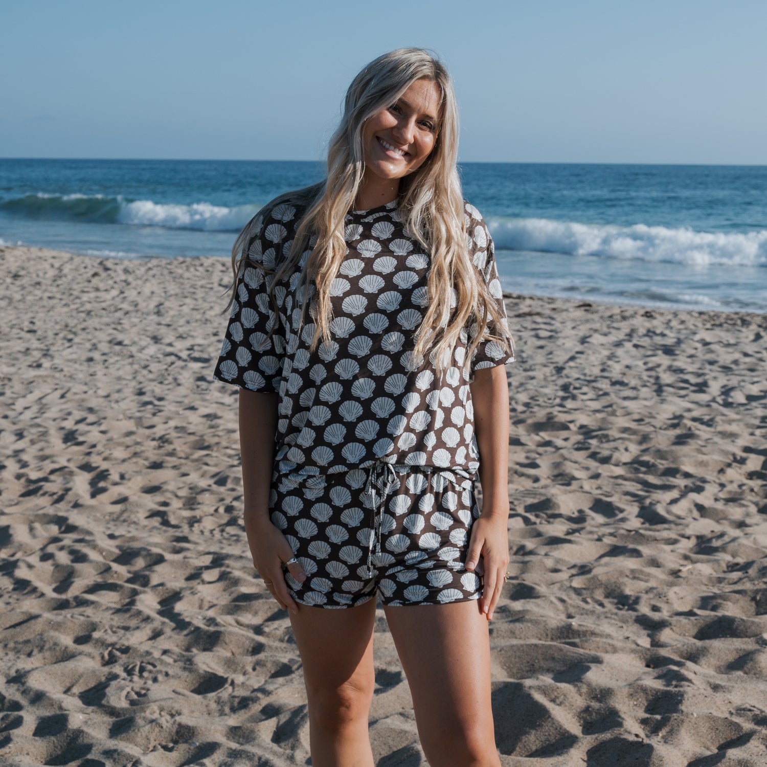 A woman with long blonde hair smiles on a sandy beach near the ocean, wearing the forever french baby Women's Bamboo Short Set | Seashells—breathable shorts and a short-sleeve top with a white seashell pattern on a dark background.