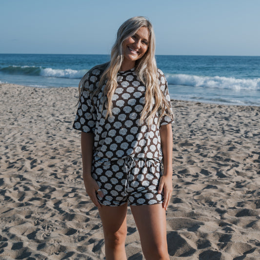 A woman with long blonde hair smiles on a sandy beach near the ocean, wearing the forever french baby Women's Bamboo Short Set | Seashells—breathable shorts and a short-sleeve top with a white seashell pattern on a dark background.
