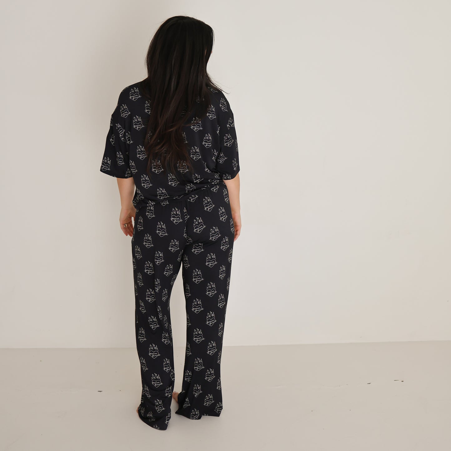 A woman with long dark hair, wearing the Women's Boxy Tee Lounge Set in Flaming Hearts by forever french baby, stands facing away from the camera in a plain white-walled room.