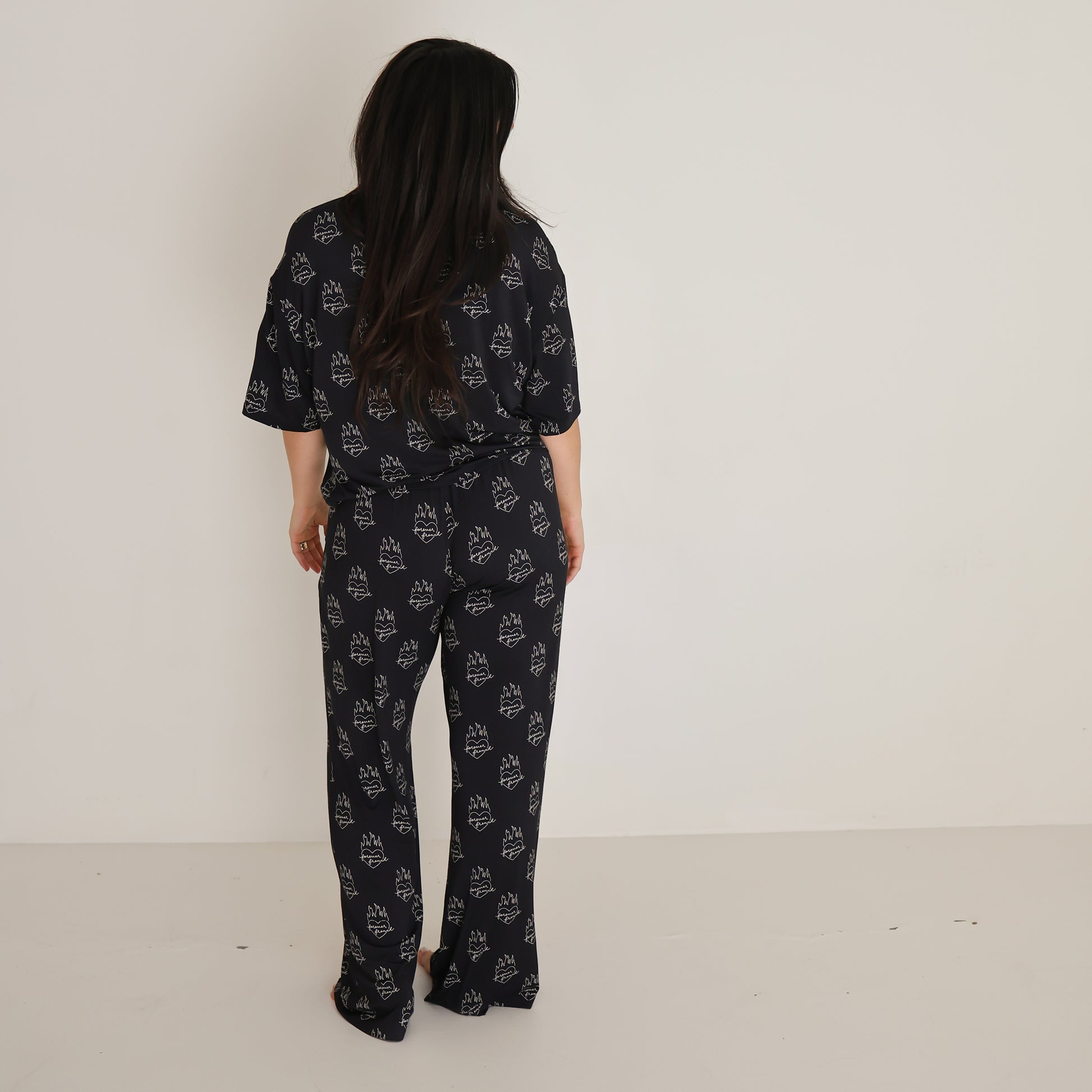 A woman with long dark hair, wearing the Women's Boxy Tee Lounge Set in Flaming Hearts by forever french baby, stands facing away from the camera in a plain white-walled room.