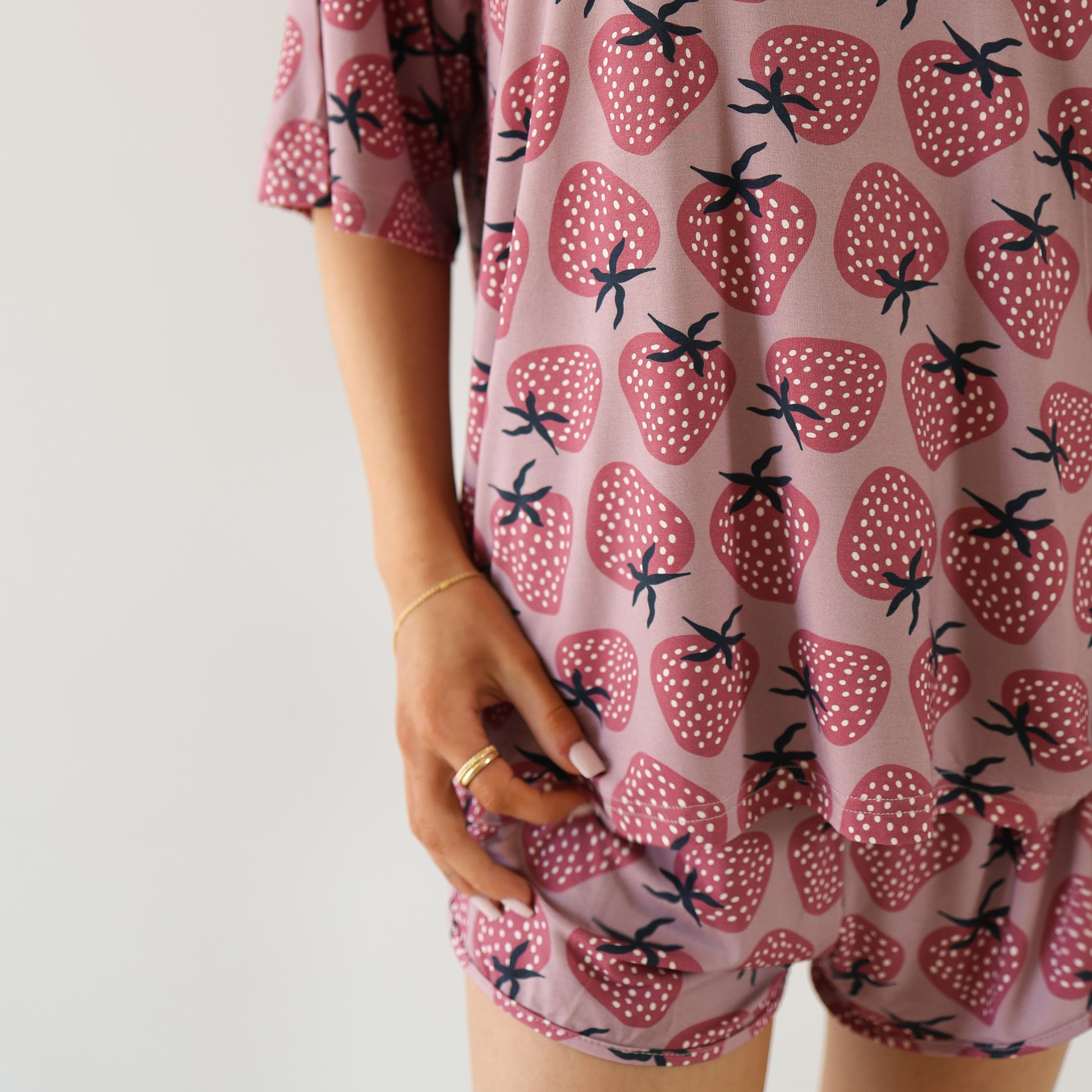 A person models the Women's Bamboo Short Set | Matilda by forever french baby, featuring pink fabric with red strawberries. Only the lower face and body are visible, standing against a white background while holding the hem of the soft shorts.
