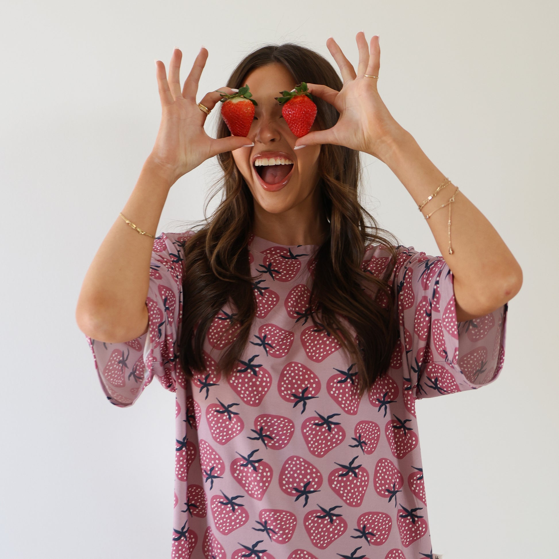 A smiling woman in the Women's Bamboo Short Set | Matilda by forever french baby, featuring a pink strawberry-patterned bamboo outfit, playfully holds two strawberries over her eyes against a plain white background.