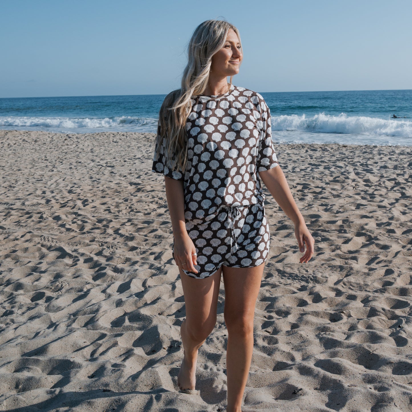 A woman with long blonde hair walks barefoot on a sandy beach, smiling in the forever french baby Women's Bamboo Short Set | Seashells. The matching loungewear features a polka dot shirt and shorts as ocean waves shimmer under a clear blue sky.
