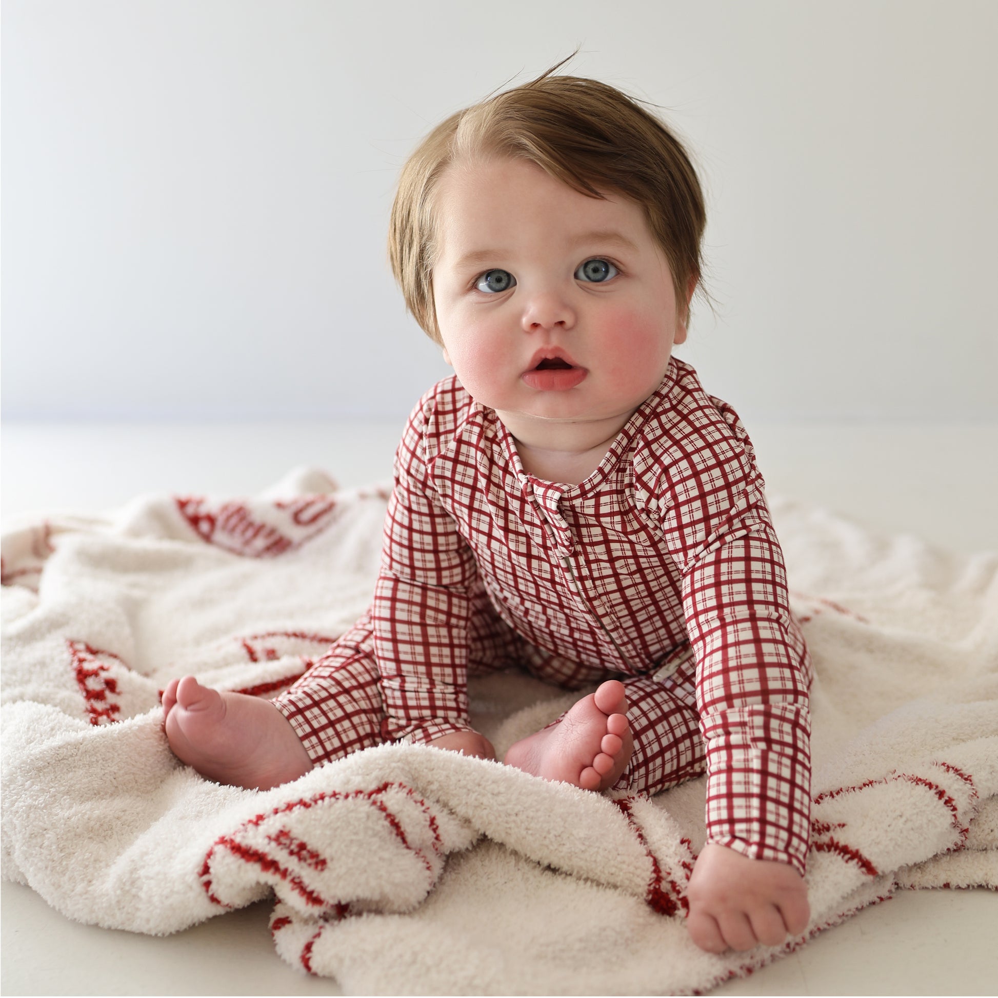 A light-skinned baby with blue eyes and brown hair sits on a soft blanket, wearing forever french baby Bamboo Zip Pajamas in the red and white Window Pane pattern, gazing upward in a bright, minimal room.