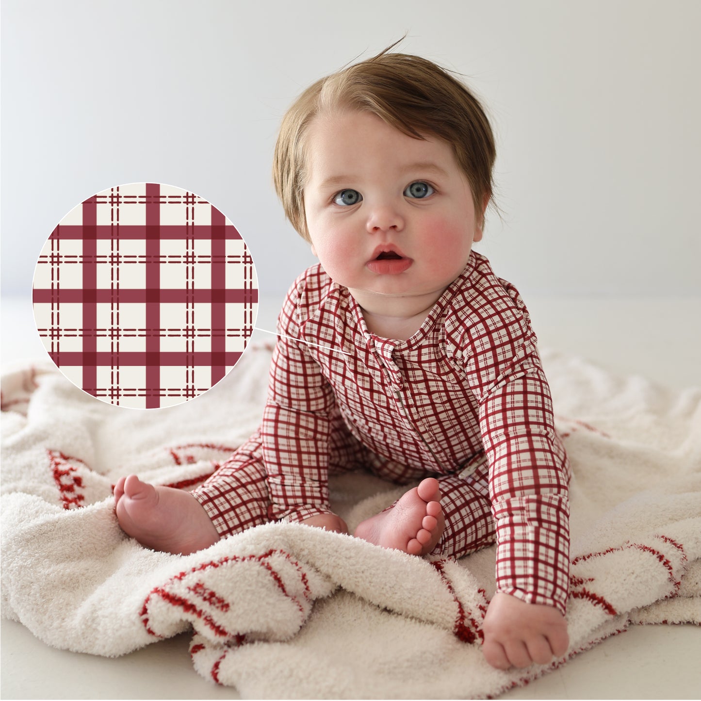 A baby sits on a soft, cream blanket with red accents, wearing forever french baby Bamboo Zip Pajamas in the Window Pane pattern. An inset features a close-up of the hypoallergenic sleepwear’s red and white plaid design.