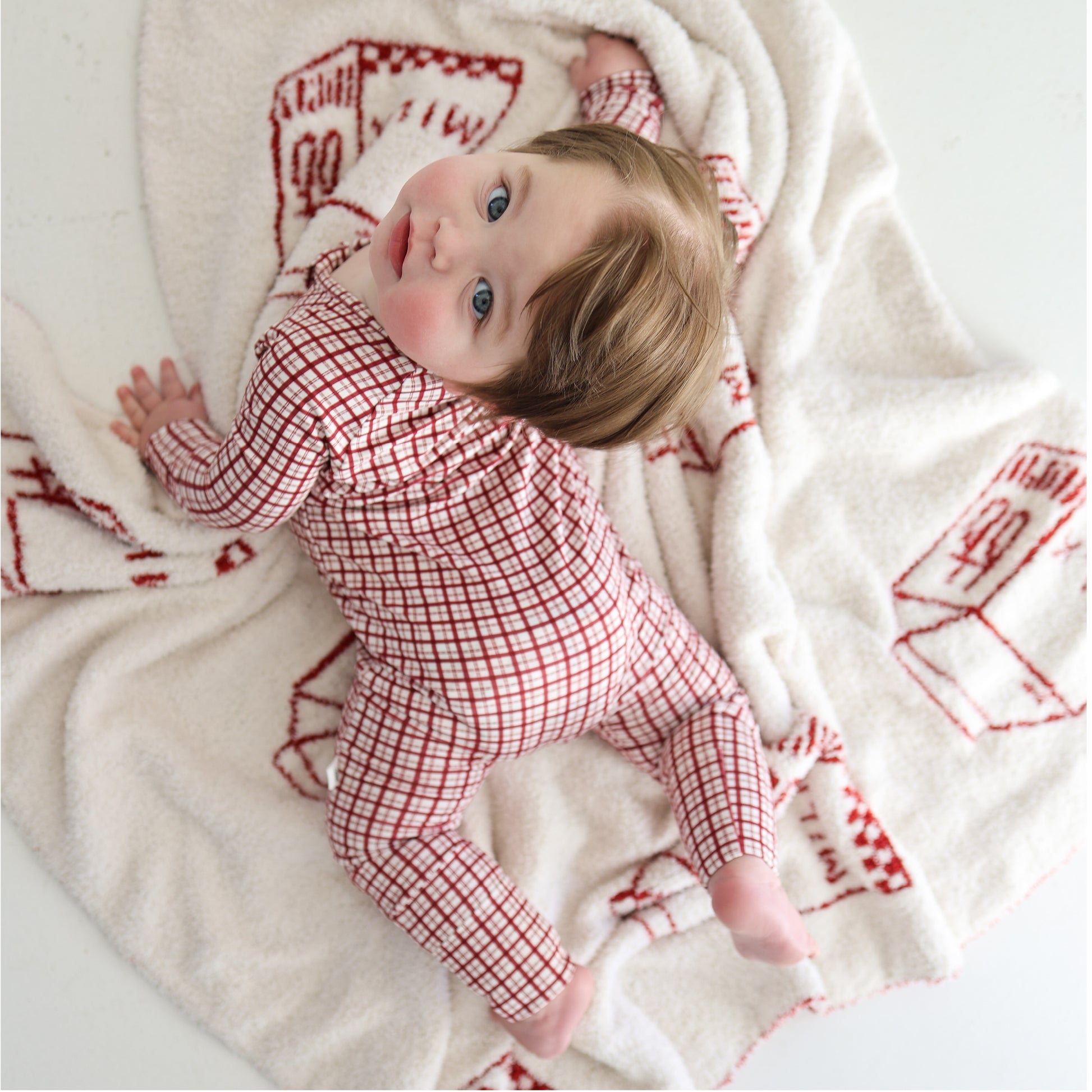 A baby wearing forever french baby Bamboo Zip Pajamas | Window Pane lies on a white blanket with red designs, looking up at the camera. The breathable pajamas highlight the baby's light brown hair and blue eyes.