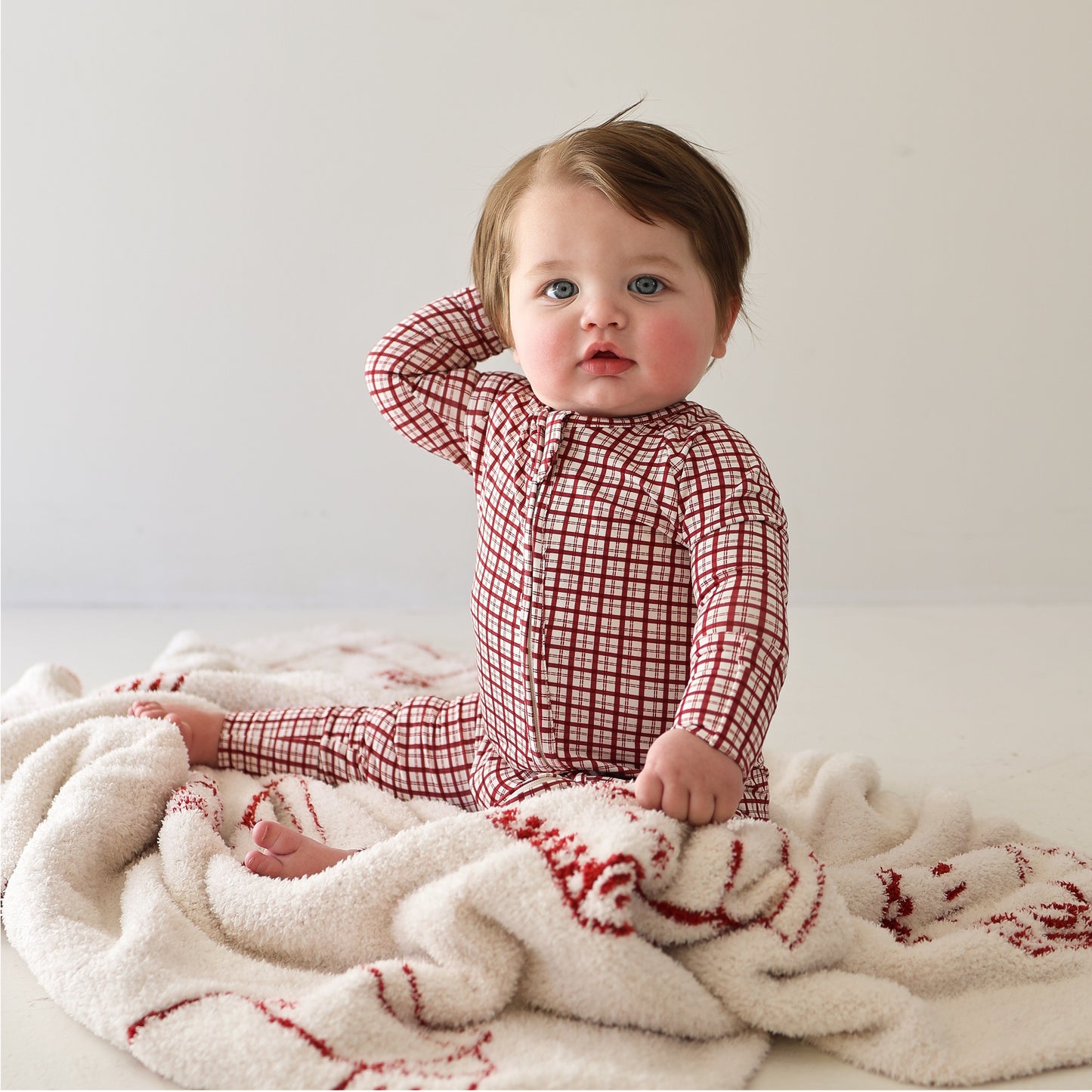 A baby in forever french baby's Bamboo Zip Pajamas | Window Pane sits on a cream-colored blanket, gazing ahead with one hand behind their head. The red and white checkered onesie stands out against the plain, light background.