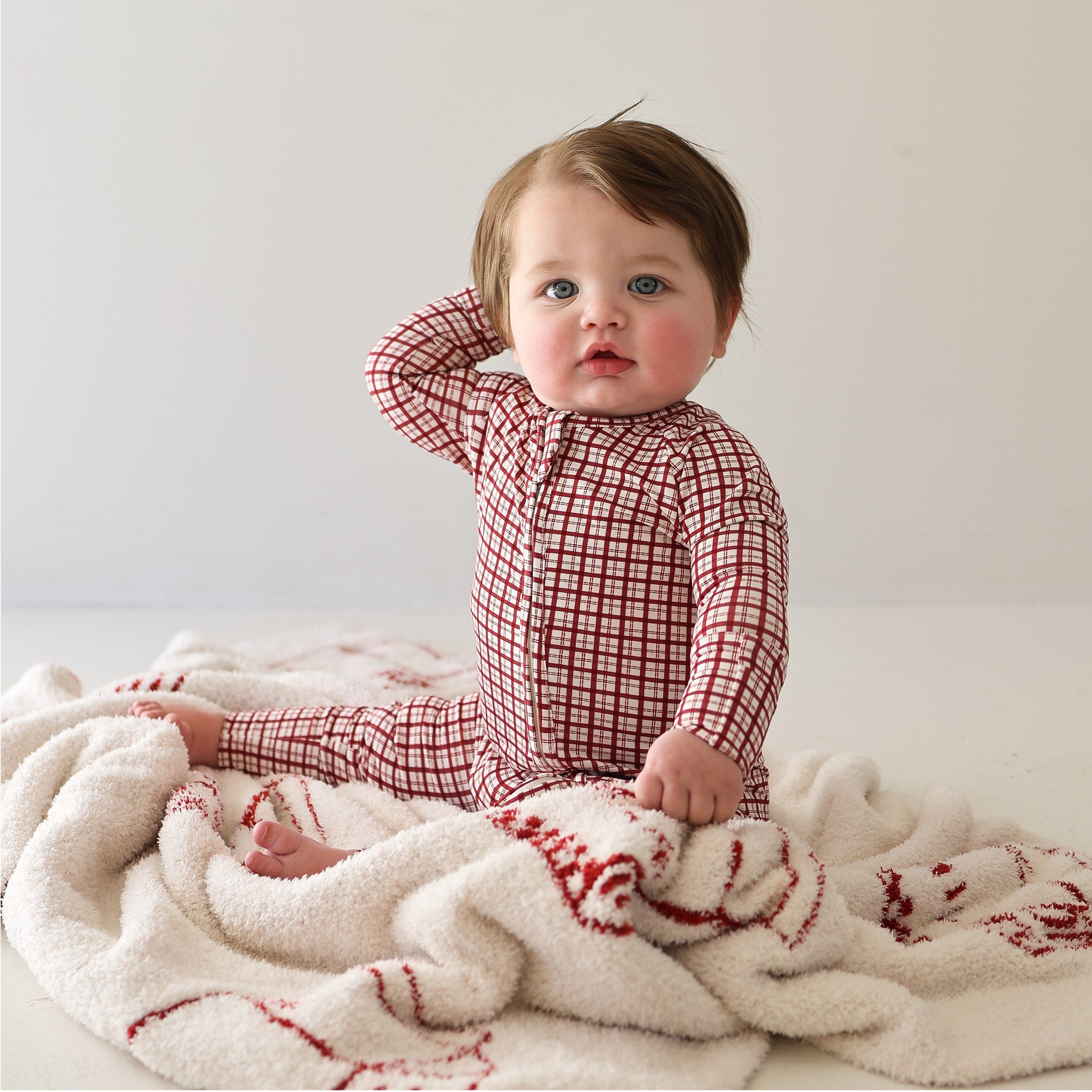 A baby in forever french baby's Bamboo Zip Pajamas | Window Pane sits on a cream-colored blanket, gazing ahead with one hand behind their head. The red and white checkered onesie stands out against the plain, light background.