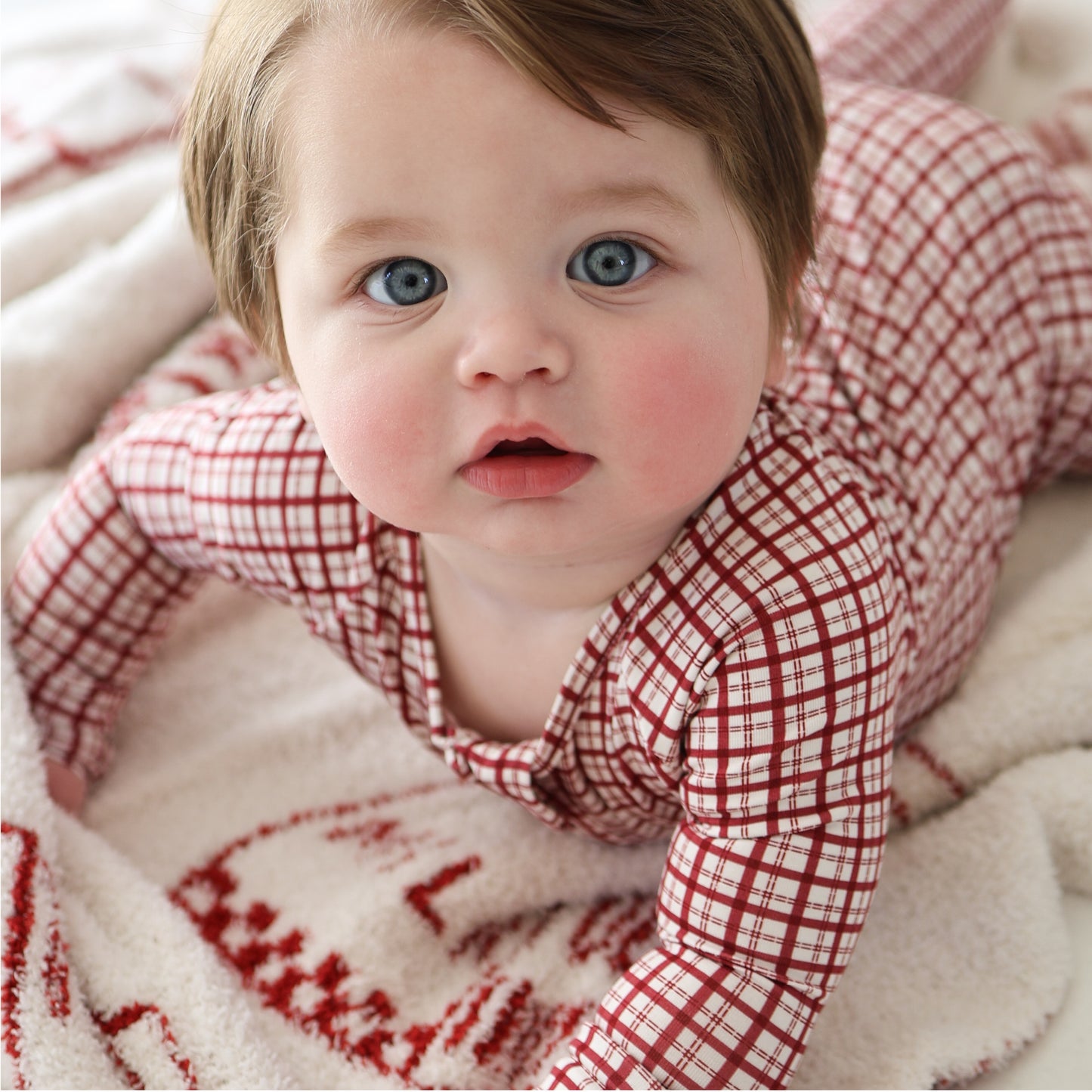 A blue-eyed, rosy-cheeked baby in forever french baby’s Bamboo Zip Pajamas | Window Pane, featuring a red and white plaid design, lies on a soft white blanket with red patterns, gazing up at the camera.