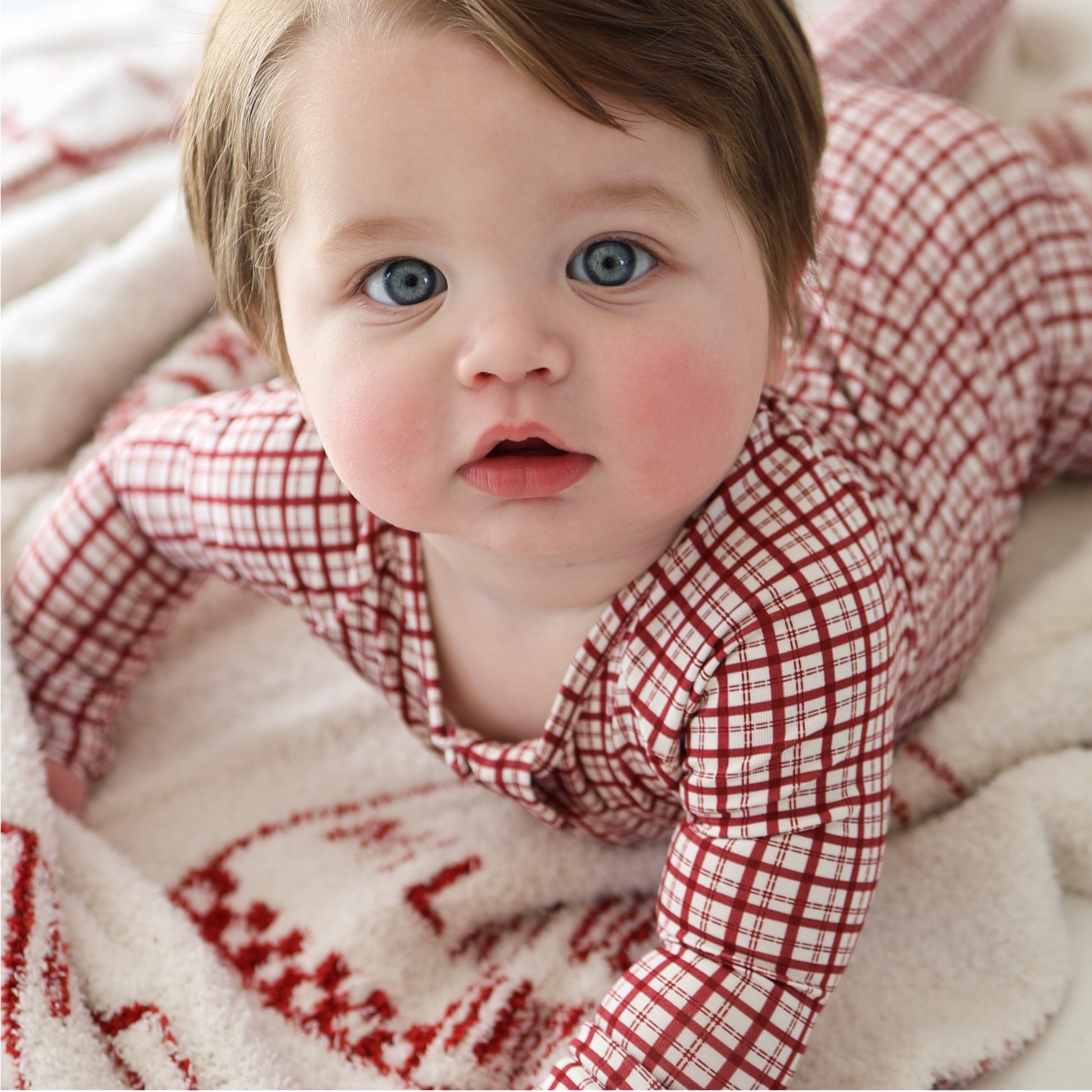 A blue-eyed, rosy-cheeked baby in forever french baby’s Bamboo Zip Pajamas | Window Pane, featuring a red and white plaid design, lies on a soft white blanket with red patterns, gazing up at the camera.