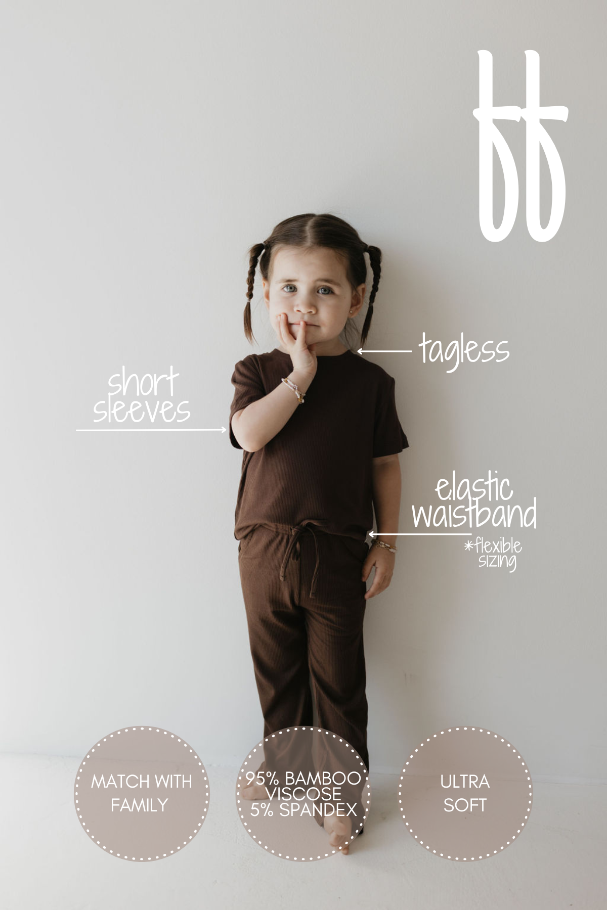 A young child with braided hair stands against a light background wearing the Toddler Short Sleeve Bamboo Lounge Set in Coffee Bean by forever french baby. This brown, tagless outfit features short sleeves and an elastic waistband and is crafted from 95% bamboo viscose and 5% spandex for an ultra-soft, breathable feel. It's hypo-allergenic and ideal for matching with the family.