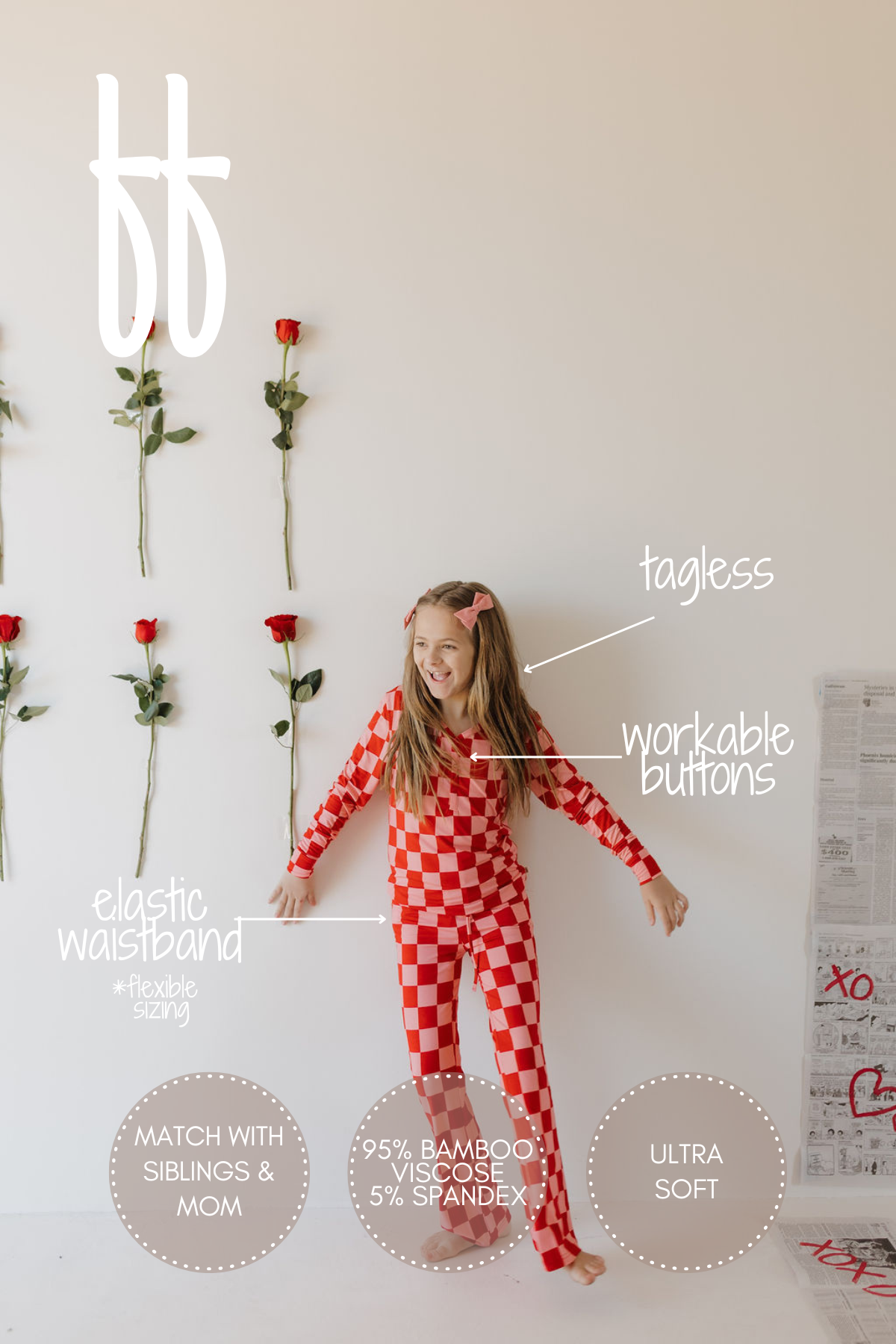 A joyful child poses in front of a white wall with red roses wearing the Youth Flare Bamboo Pajamas | XOXO by forever french baby. The red and white checkered onesie is made from breathable, hypo-allergenic fabric (95% bamboo viscose, 5% spandex) featuring an elastic waistband and tagless design.