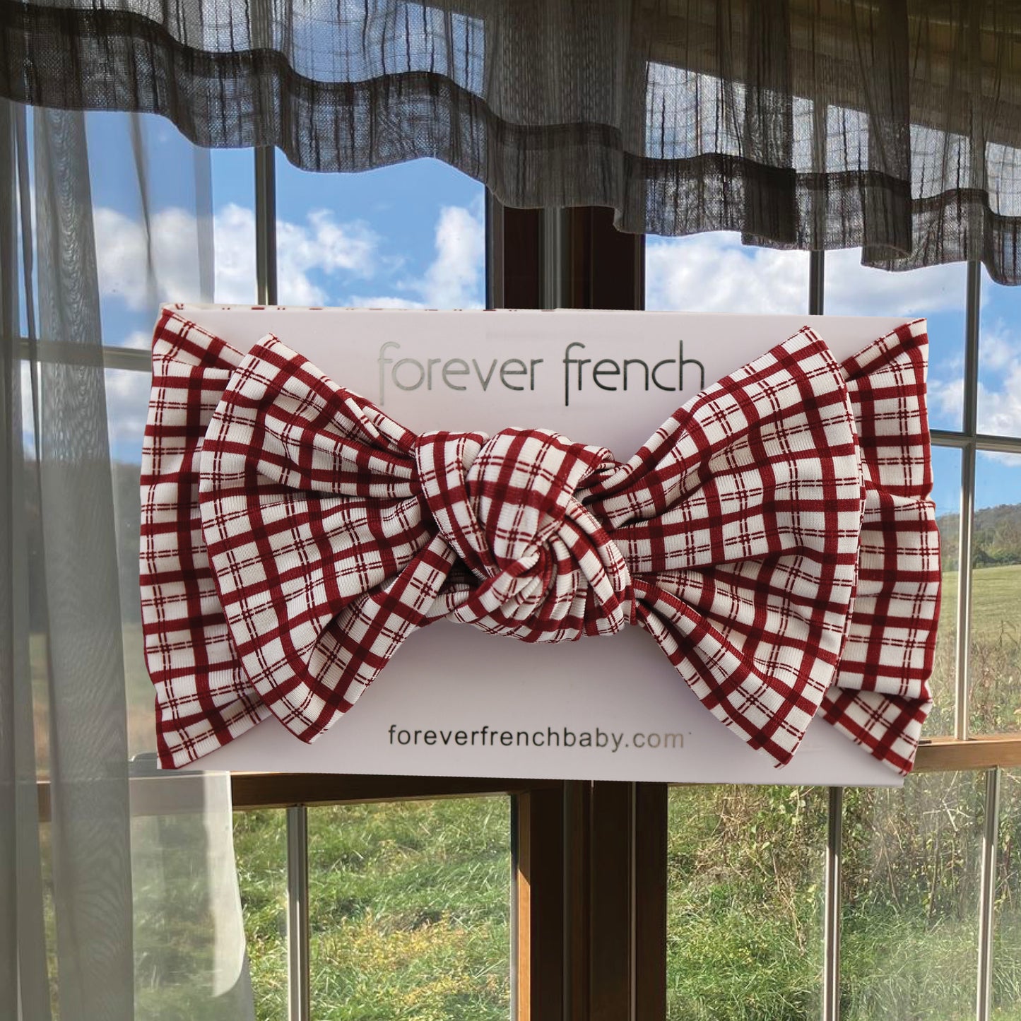 A Bamboo Head Wrap in a red and white window pane pattern from forever french baby is displayed on a branded card in front of a window with sheer curtains, showcasing green grass and blue sky with clouds outside.