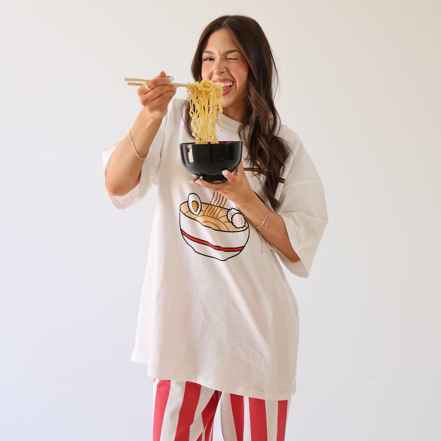 A woman in a white oversized ramen graphic T-shirt and forever french baby Women's Wide Leg Pants | Tokyo Stripe smiles while eating noodles with chopsticks from a black bowl, standing against a plain white background.