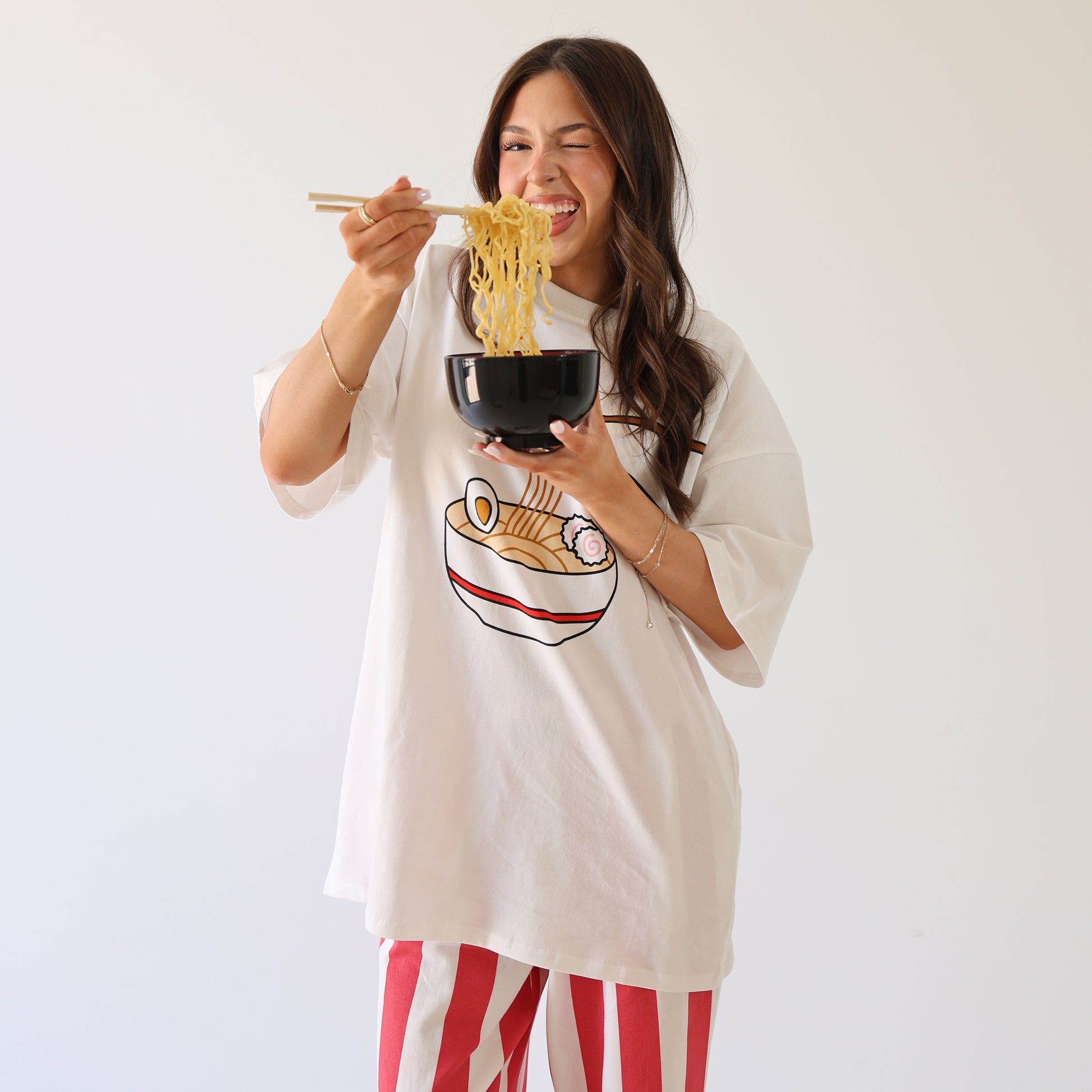 A woman in a white oversized ramen graphic T-shirt and forever french baby Women's Wide Leg Pants | Tokyo Stripe smiles while eating noodles with chopsticks from a black bowl, standing against a plain white background.