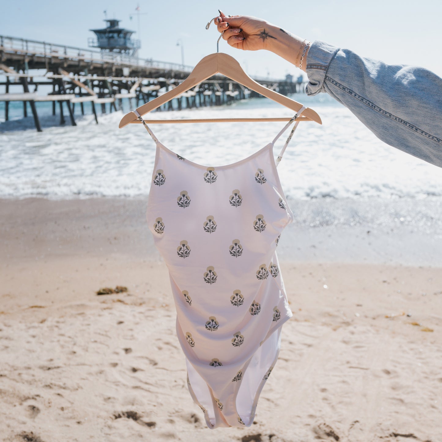 A person holds a hanger with a Forever French Women's One Piece Swimsuit | Marigold on a sandy beach, as ocean waves and a wooden pier appear in the background beneath a bright sky.