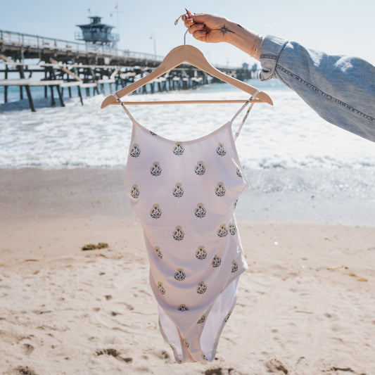 A person holds a hanger with a Forever French Women's One Piece Swimsuit | Marigold on a sandy beach, as ocean waves and a wooden pier appear in the background beneath a bright sky.