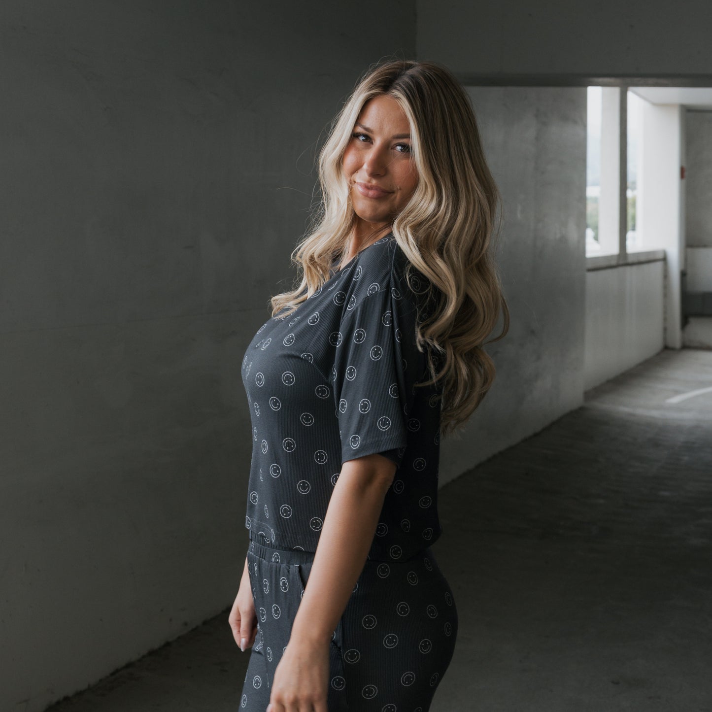 A woman with long, wavy blonde hair smiles slightly in a parking garage, wearing the forever french baby Women's Boxy Tee Lounge Set | Smiley Ribbed—a matching dark lounge set with small circular patterns made from hypo-allergenic bamboo.