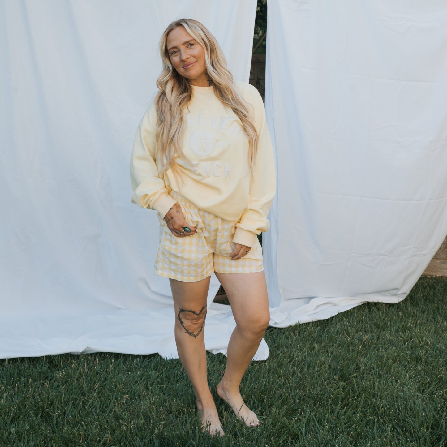 A woman with long blonde hair, barefoot on grass, wears the forever french baby Adult Sweatshirt | ff Yellow and yellow gingham shorts. She smiles softly, showing a heart tattoo on her thigh. White sheets hang in the background.
