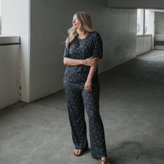 A woman with long blonde hair smiles in a parking garage, wearing the forever french baby Women's Boxy Tee Lounge Set in Smiley Ribbed and comfy sandals, with one arm held gently.