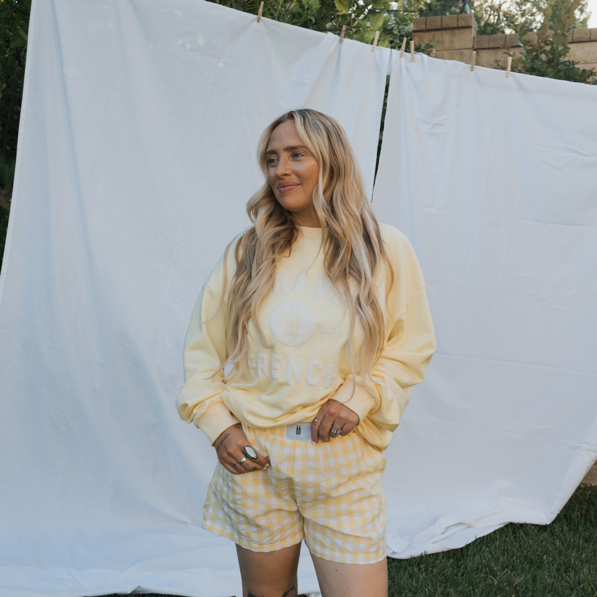 A woman with long blonde hair stands on grass, smiling softly in a forever french baby Adult Sweatshirt | ff Yellow. She pairs it with yellow gingham shorts, with a white sheet backdrop—perfect for everyday wear.