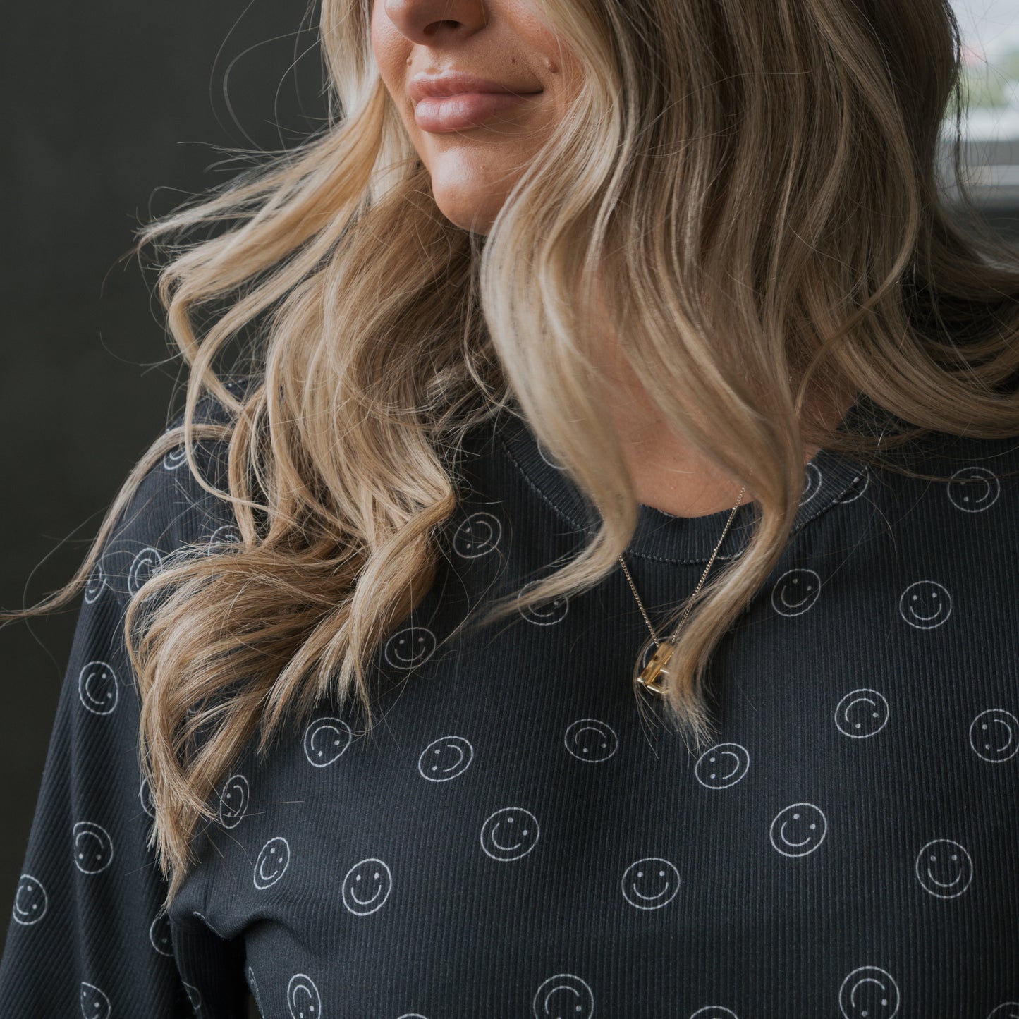 A woman with long, wavy blonde hair wears the Women's Boxy Tee Lounge Set | Smiley Ribbed from forever french baby. The black ribbed set features white smiley faces. She pairs it with a gold necklace and smiles softly, her face partly visible.