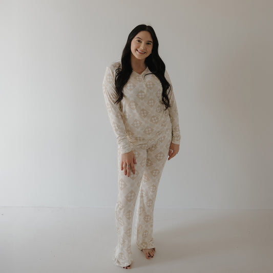 A woman with long dark hair stands barefoot in a minimalist room, smiling and wearing the forever french baby Women's Long Sleeve Bamboo Pajama | Cottage Quilt—a light, patterned zip-up top and loose pants set for comfort and style.