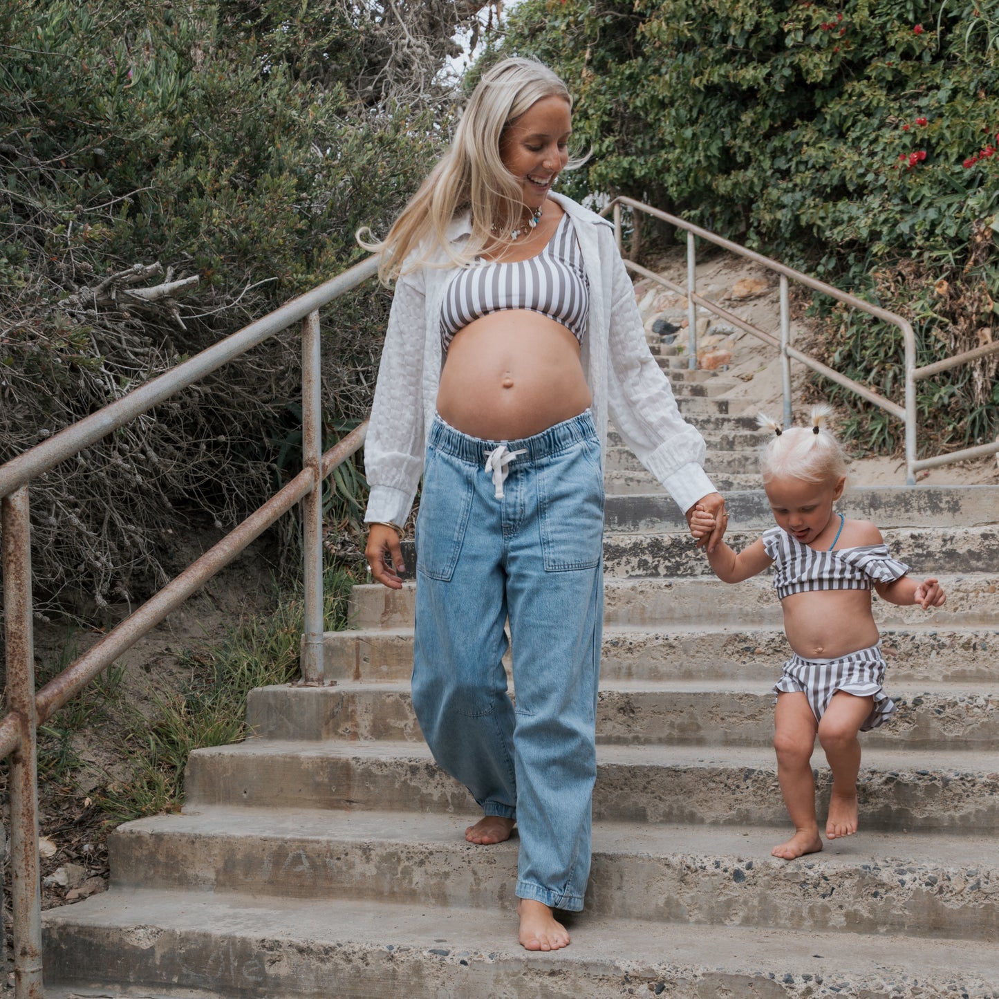 A woman and a young girl, both barefoot and wearing forever french baby's Women's Two Piece Swimsuit in Olive Stripe, walk hand-in-hand down stone steps surrounded by greenery. The woman gently guides the girl as they descend together.