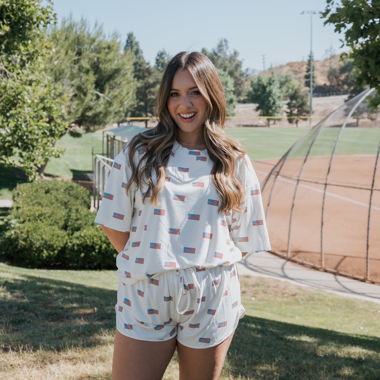 A woman with long brown hair smiles while standing on grass near a baseball field, wearing the forever french baby Women's Bamboo Short Set | Americana Ribbed. Small American flag patterns decorate the matching set. Trees and the field appear in the background.
