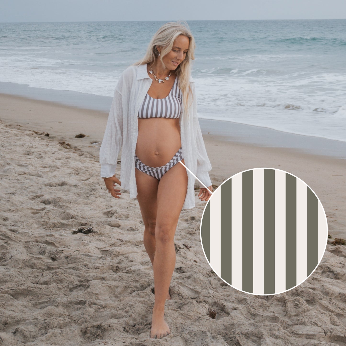 A pregnant woman in the Women's Two Piece Swimsuit | Olive Stripe by forever french baby, featuring gray and white vertical stripes, walks barefoot on a sandy beach with the ocean in the background.