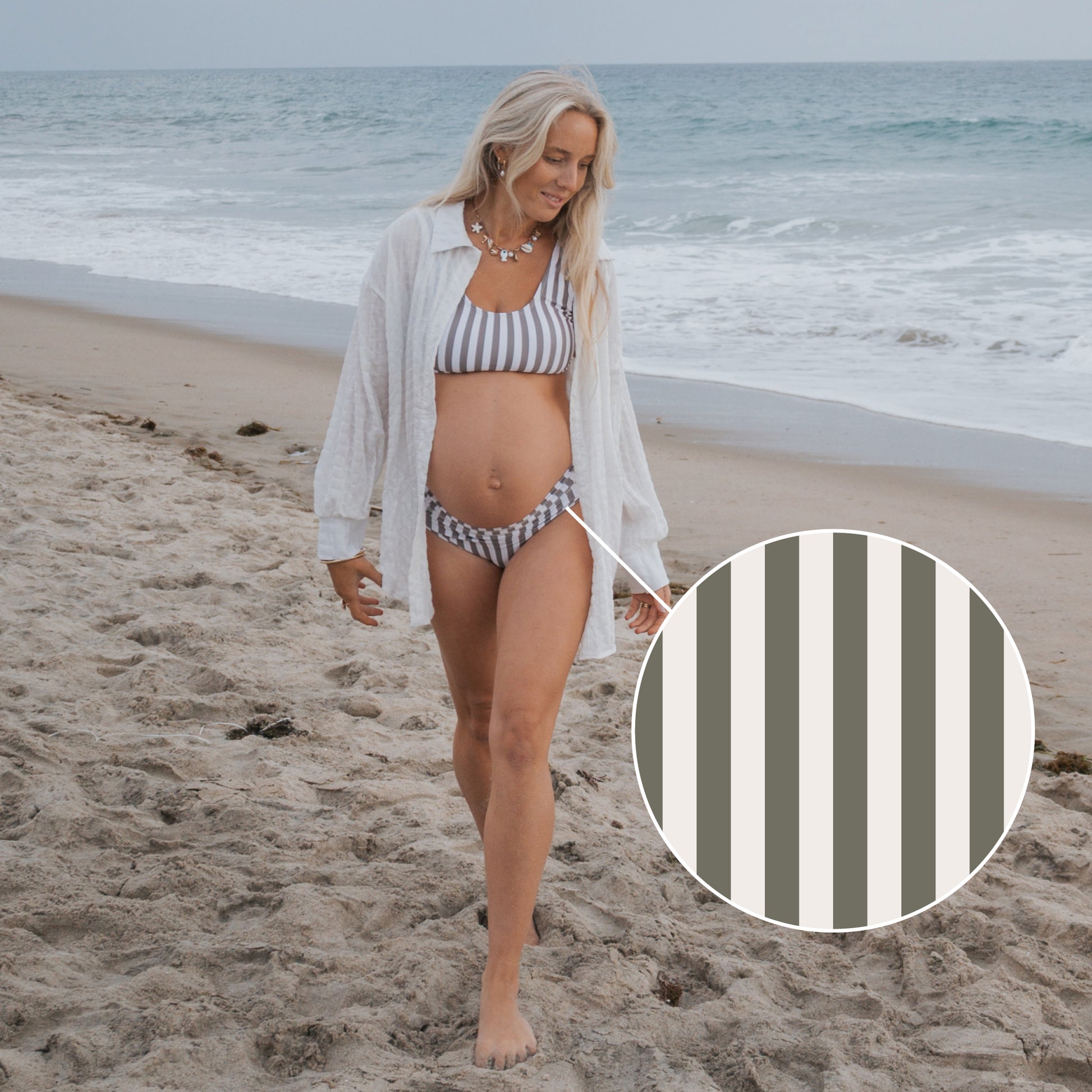 A pregnant woman in the Women's Two Piece Swimsuit | Olive Stripe by forever french baby, featuring gray and white vertical stripes, walks barefoot on a sandy beach with the ocean in the background.
