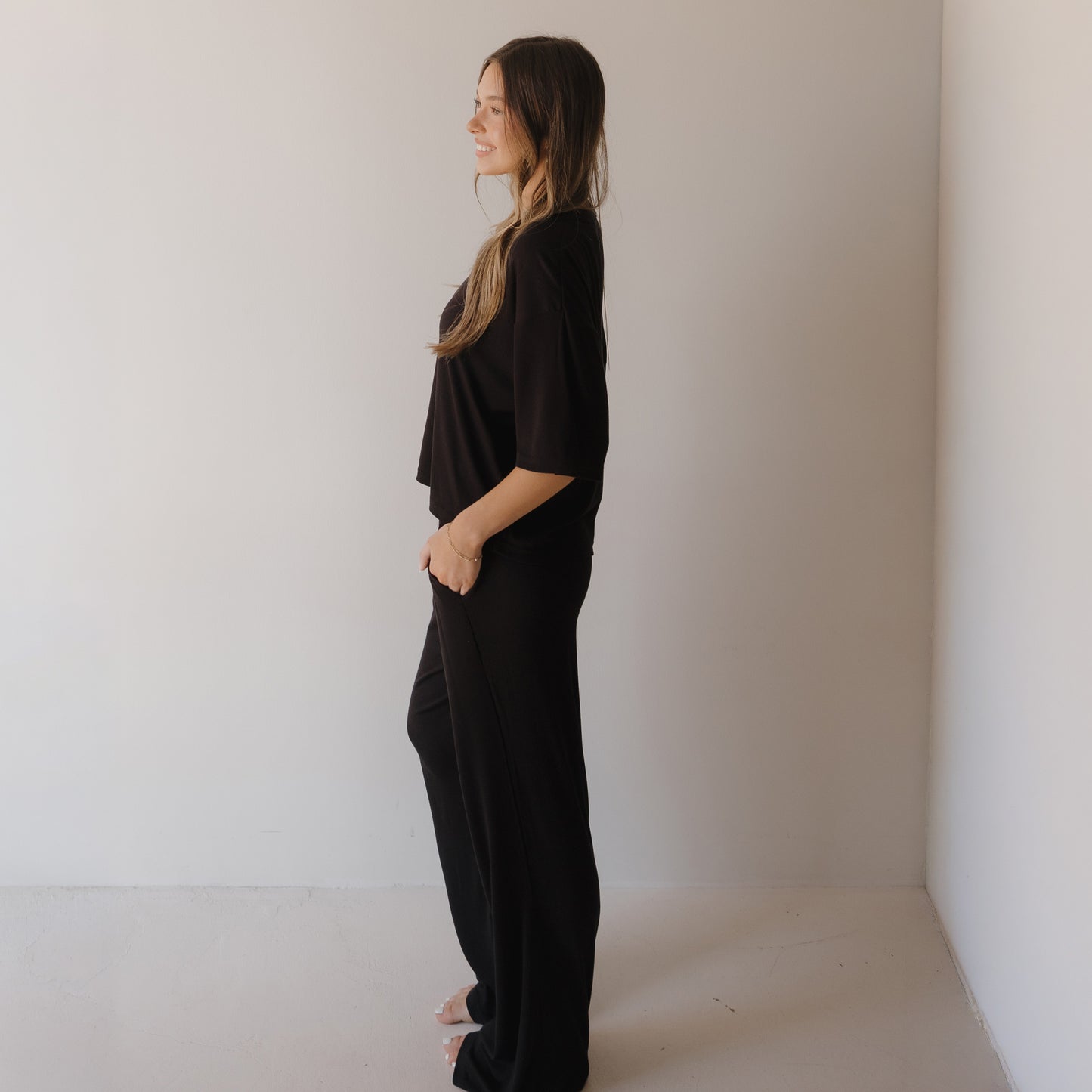 A woman with long hair stands in profile, smiling in a minimal, light-toned room as she wears the forever french baby Women's Boxy Tee Lounge Set | Ribbed Black.