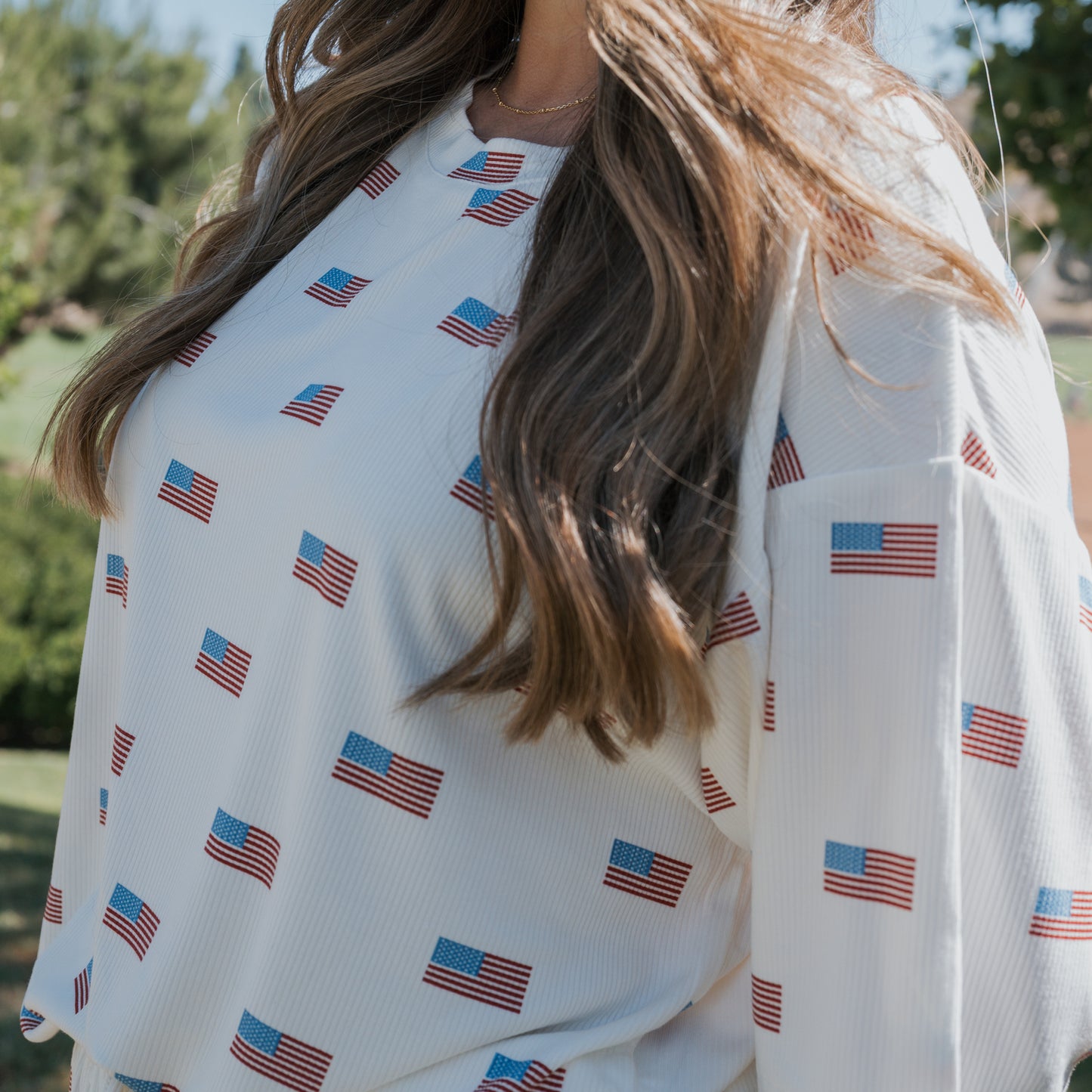 A woman with long brown hair wears the forever french baby Women's Bamboo Short Set | Americana Ribbed, featuring a white shirt with small American flag patterns, made from breathable loungewear fabric, while standing outdoors amid greenery.