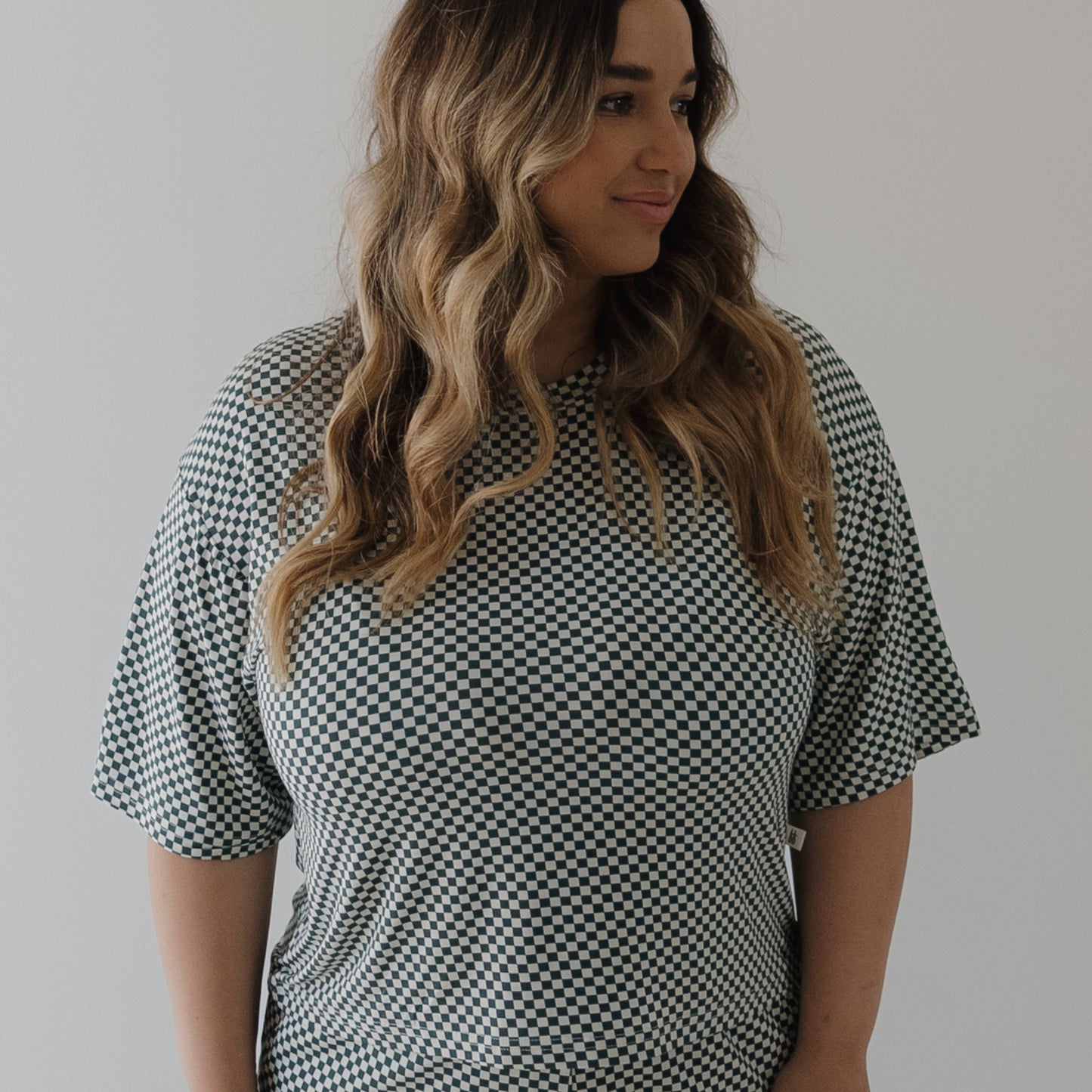 A woman with long wavy hair models the forever french baby Women's Boxy Tee Lounge Set in Lucky Micro Check, standing against a light background and smiling softly as she looks slightly to her right.