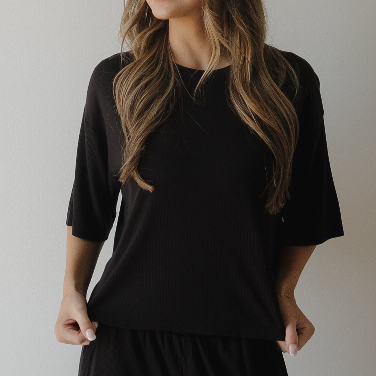 A woman with long wavy hair poses against a plain light background, holding the sides of her shirt. She wears the forever french baby Women's Boxy Tee Lounge Set in Ribbed Black, and her face is partially out of view.